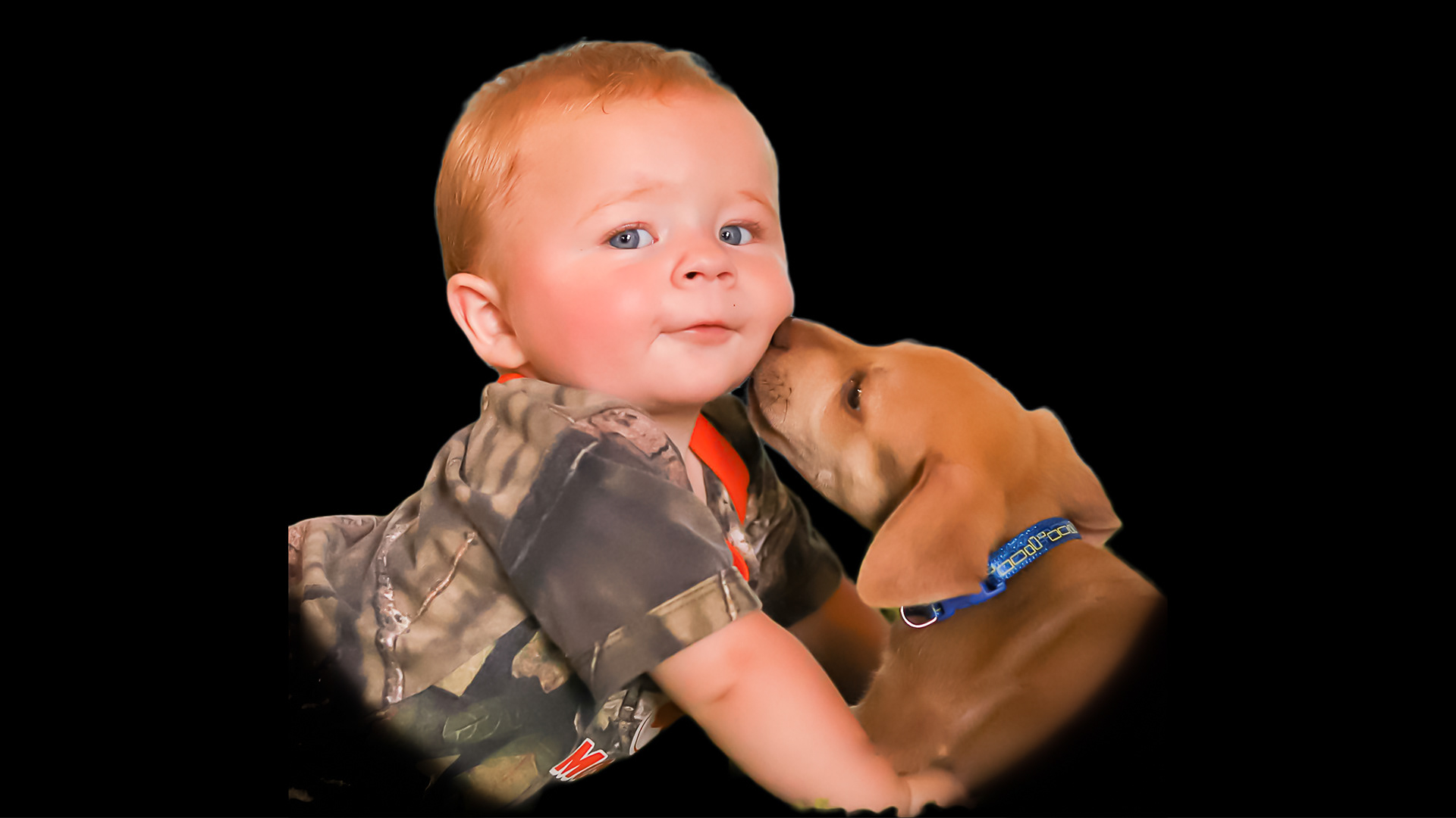A small reddish brown colored puppy kissies a reddish brown haired baby boy on the cheek.