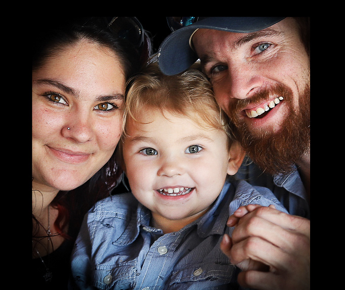 A family of 3 smiles while standing cheek to cheek.