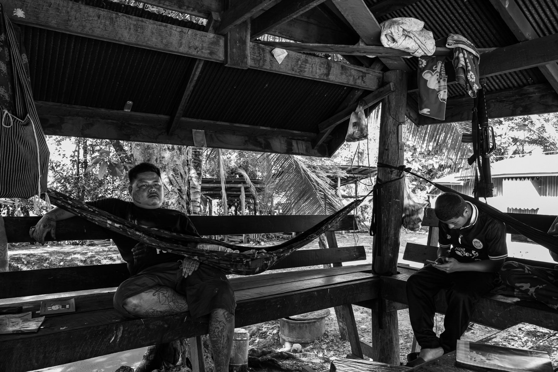 Two Karen National Liberation Army soldiers sit inside a simple shelter, with one weapon visible, taking a moment of rest amid ongoing clashes in Karen State.  Wale, Karen State, Myanmar, October 2025