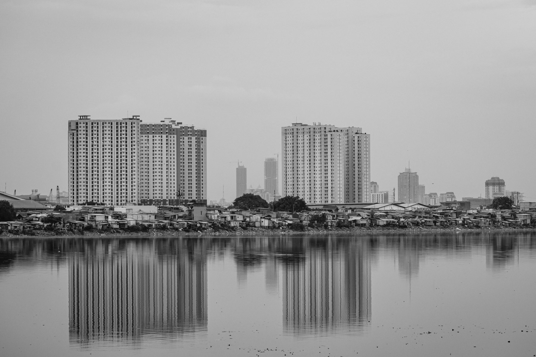 Jakarta the Indonesian capital of more than 10 million is sinking! As rising sea levels were not bad enough, the capital also suffers of severe subsidence or surface sinking. Its buildings and population extract most of their water needs by drilling water wells into the underground aquifers, rendering it susceptible for collapse. Experts estimate that by 2050 more than 95% northern Jakarta's coastal districts could be submerged. Also, one of the main reasons of the increasing scale of floods in Jakarta is because the city is sinking and that rain water tends to stagnate rather than to wash out to the sea as coastal areas where rivers flow out are under the sea level. During the past decennia river water became too polluted to be used as the main source for piped water, and a shift was made towards digging deep water wells all over the city. This happened in an un-controlled and often illigal way, draining drop by drop the underground aquifers on which the city rests and creating enormeous airpockets making the surface unstable and eventually making the ground to sink and collapse. Almost half of Jakarta lies now below sea level and coastal districts of the city are sinking at an alarming rate of up to 25cm per year! Floodings, subsidence and traffic congestion are the main reasons why the Indonesian authorities want to relocate the capital to the island of Borneo in the next few years. Jakarta is sinking faster than any other mega city in the world, faster than the rise of the sea caused by global warming. One of the massive ongoing (development) projects, which the authorities say will help rescue the city of being submerged, is the "Great Garuda" in the Bay of Jakarta, consisting of a 30 Km outer sea wall along several artificial islands packed with luxary real-estate. The idea is to create and artificial lagoon in which water levels could be controlled and lowered in order to allow the city's rivers to drain naturally into the lagoon and eventually the sea. This in return would help to prevent deadly floods which are becoming a major issue. Experts and environmentalists argue that such project is not going to stop the sinking of the city for which the sole viable solution would be to stop all ground water extraction and only rely on other sources of water such as piped water from reservoirs filled with river and rain water. For this more effort should be invested in decontaminating and cleaning rivers, dams and lakes. The social impact of the city's subsidence, floods and frantic real-estate development is huge on the autochthon population living along the rivers and in the coastal districts. Neighborhoods are being nearly permanently flooded, re-claimed for real-estate projects, or partly evacuated to build concrete river embankments and sea walls, or just because the grounds become to unstable to live on them, pushing soon hundred thousands of people in an extreme precarious situation. Fishermen will in the immediate future have their livelihood compromised as the Bay of Jakarta will become clogged with artificial islands and sea wall affecting natural currents and fish stock. Muara Buru neighbourhood in North Jakarta where most of the soil is sinking at a rate of 10 to 20cm per year. The unstability of the ground is provoked by widespread drilling of water wells for new real estate projects, creating huge air pockets once the water is exhausted. Waduk Pluit, North Jakarta, Indonesia, March 2020