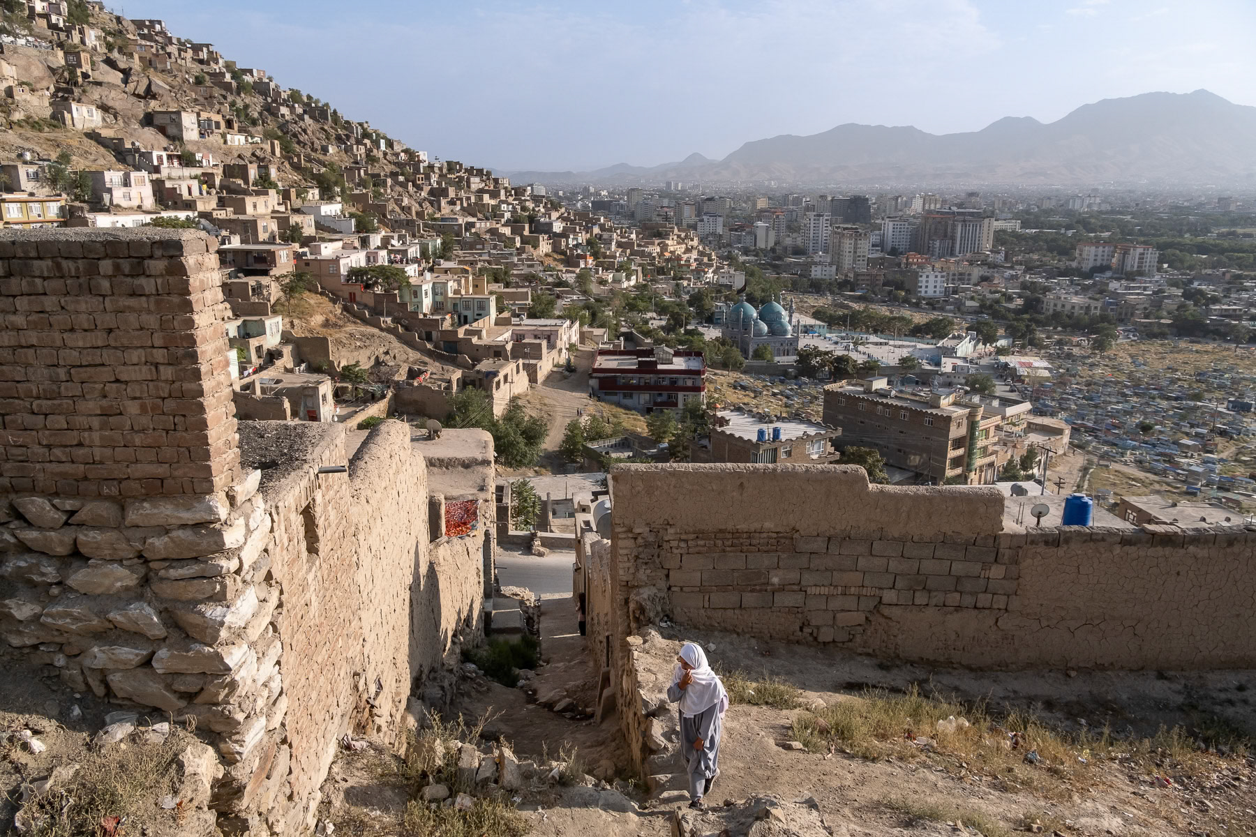 Hillside homes overlooking the urban Hazara neighbourhood of Kabul with a woman walking up the steep path, Afghanistan, August 2024