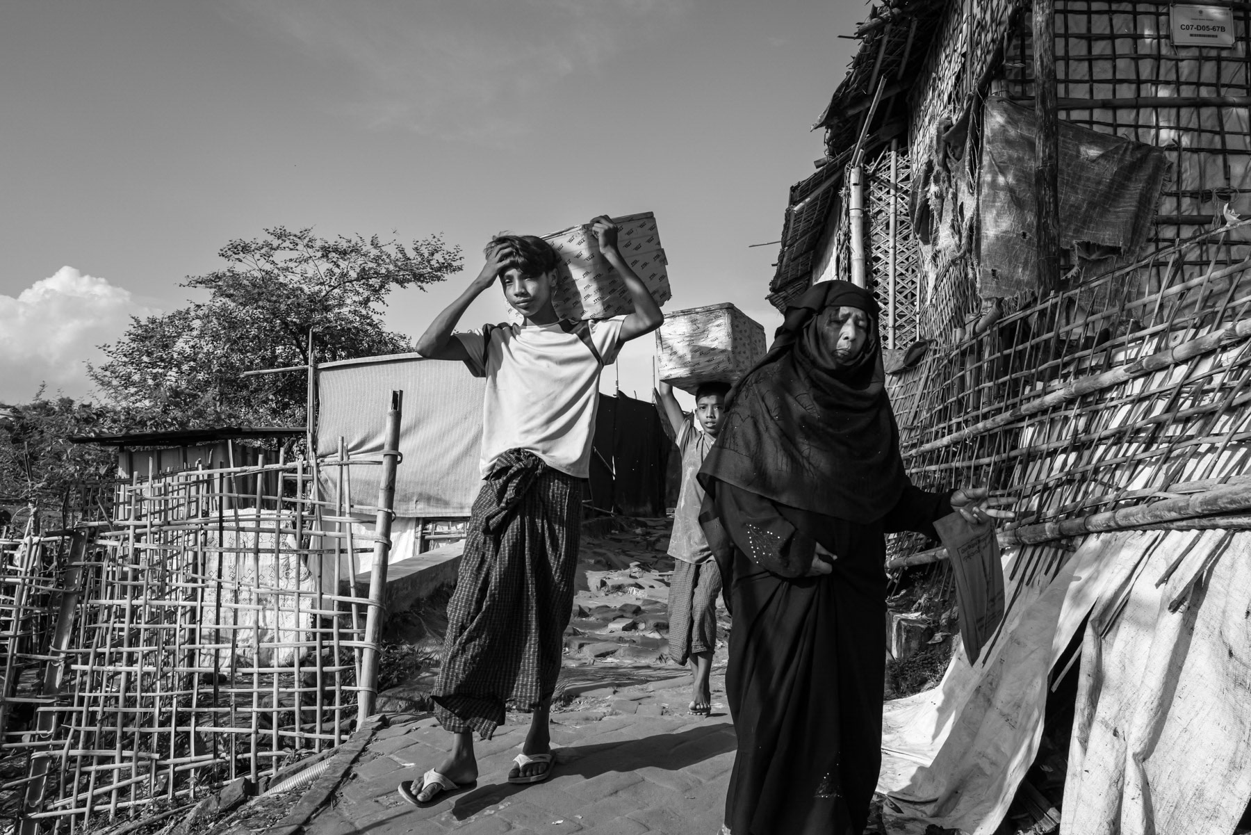 An elderly Rohingya woman carefully descends a paved alley in the camp, while two young adolescents carry goods on their heads. The settlement now hosts well over **1 million refugees**, making it the largest refugee camp in the world. Since 2024, at least **150,000 more Rohingya** have fled violence carried out by the Arakan Army (AA). Kutupalong refugee camp, Ukhiya, Bangladesh, September 2025