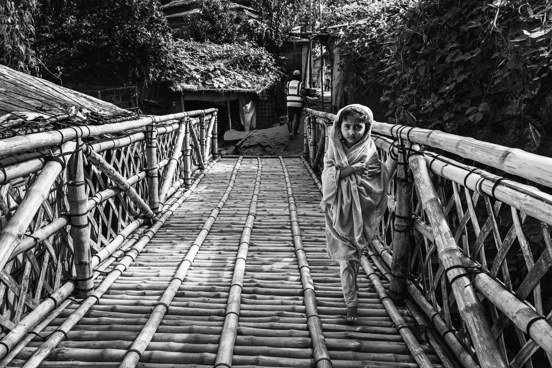 A Rohingya girl carefully crosses a bamboo bridge connecting shelters in the densely packed refugee camp. Kutupalong refugee camp, Cox's Bazar, Bangladesh, September 2025