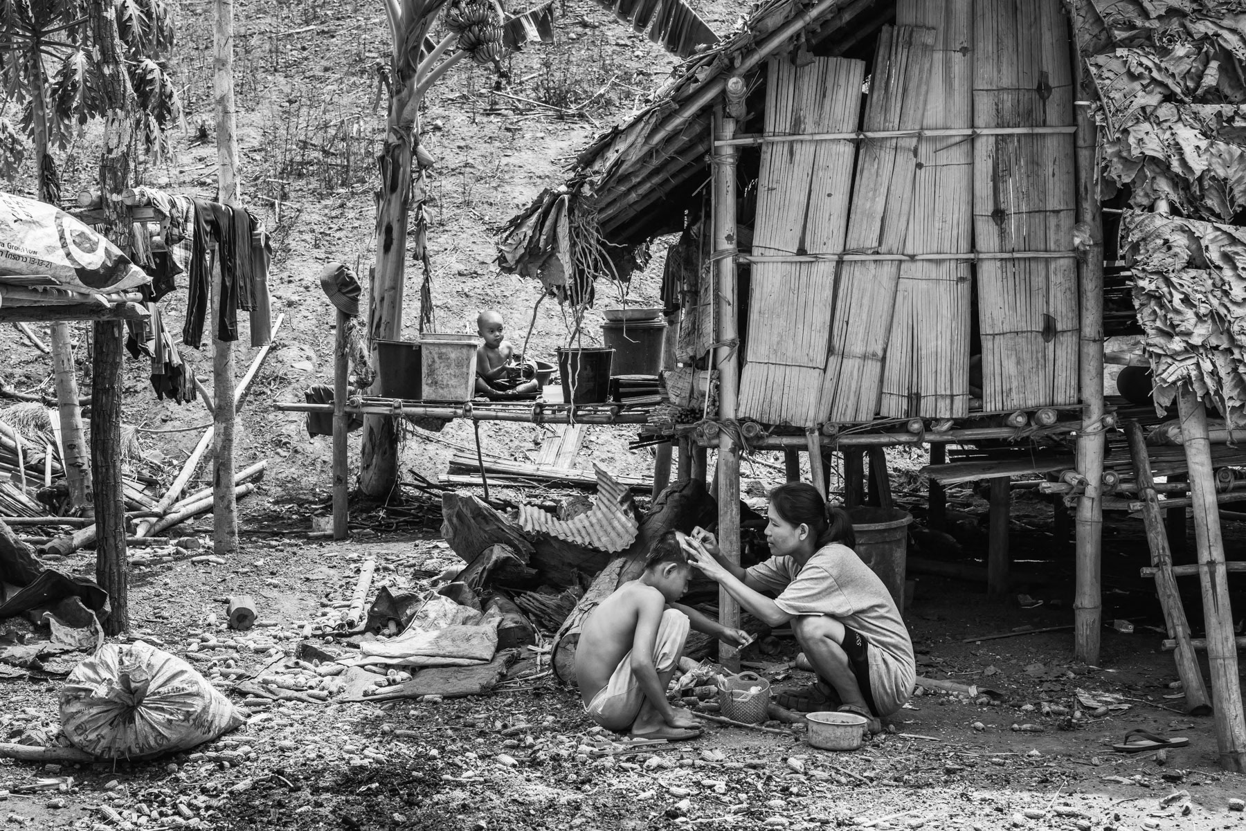Karen woman shaving her son's head as a prevention against lices. They are uprooted due to the conflict and risk of villages being hit by air strikes. Htaw IDP camp near Kanele in KNLA controlled territory. Kayin State, Myanmar, May 2024