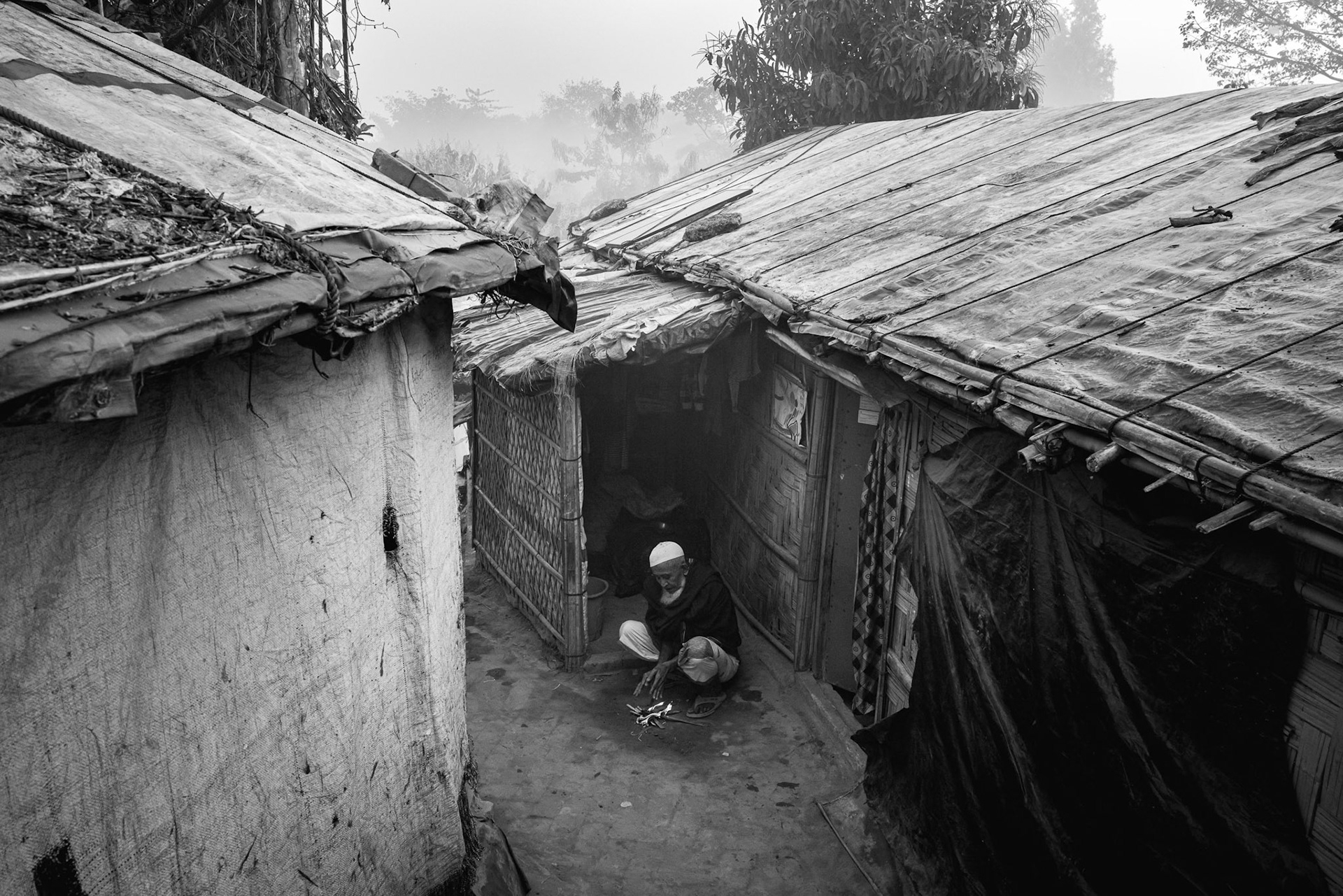 A person seated at the entrance of their home. This image captures the quiet endurance of a family that has survived extreme violence and continues to seek dignity within the confines of the world's most crowded displacement settlement. Balukhali camp, Bangladesh, January 2026