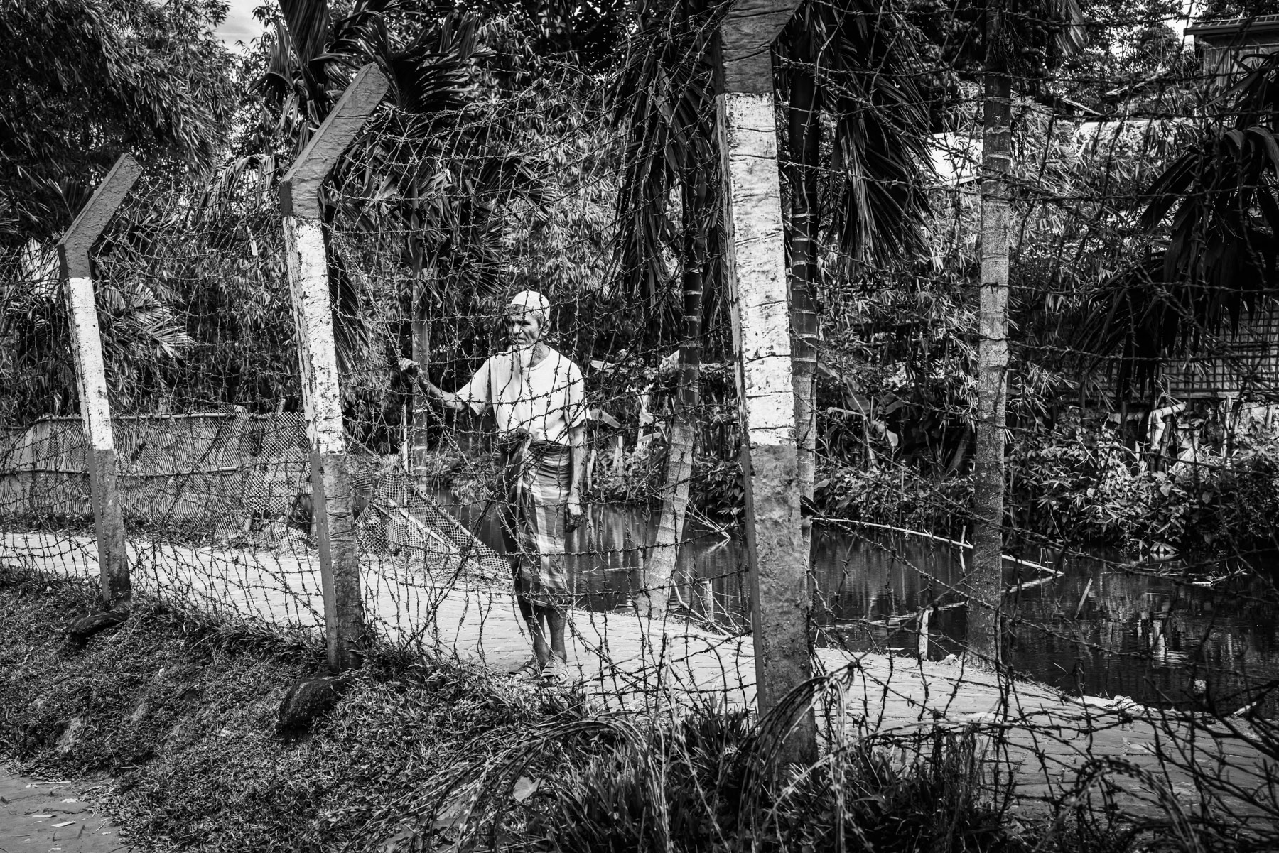 An elderly Rohingya man stands along the barbed-wire fence encircling the refugee camp, a barrier that severely restricts movement both in and out of the camp and between its different sectors. Kutupalong refugee camp, Cox's Bazar, Bangladesh, September 2025