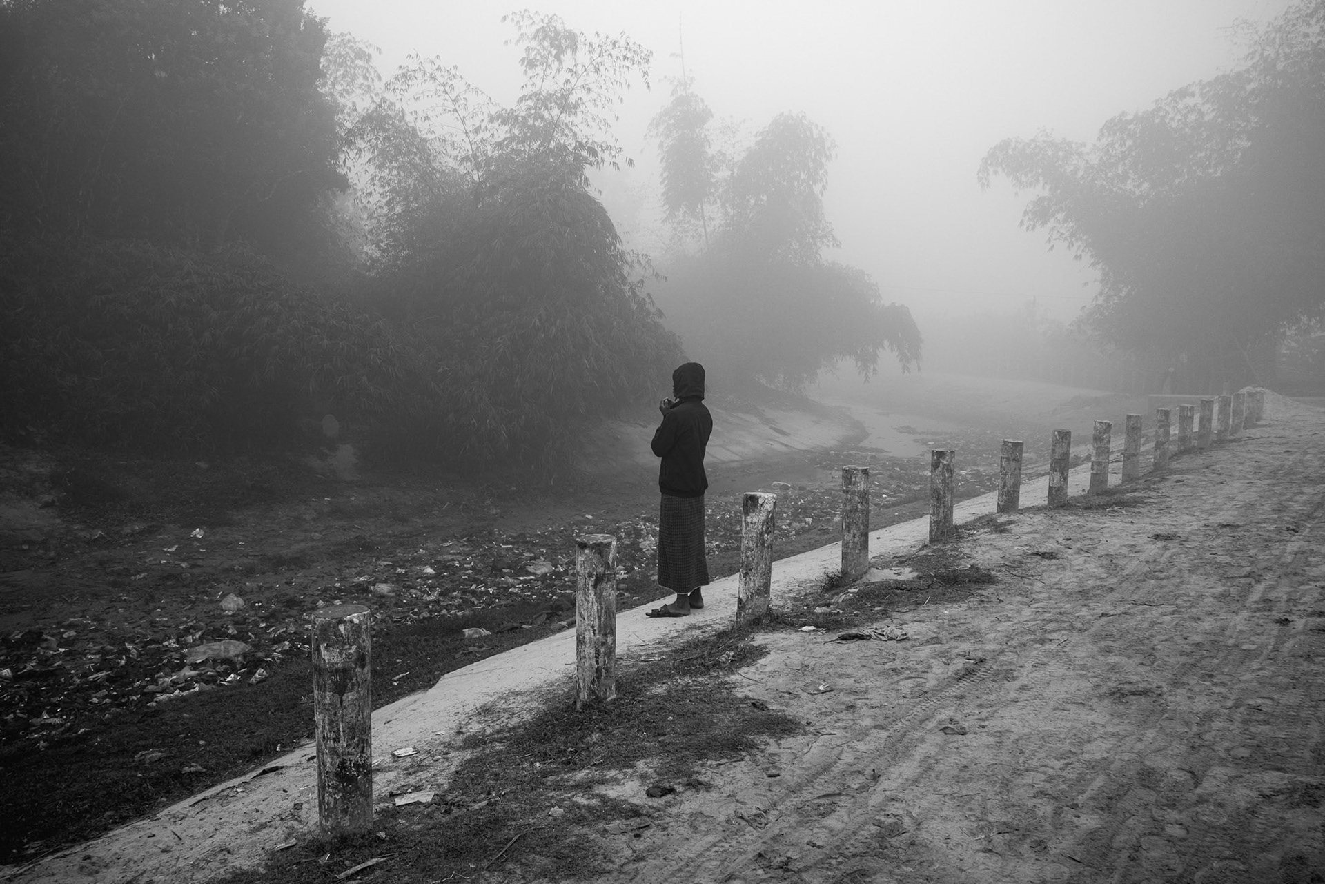 A misty morning view of a Rohingya refugee standing along a concrete-post fence overlooking a low-lying area. Balukhali camp, Bangladesh, January 2026