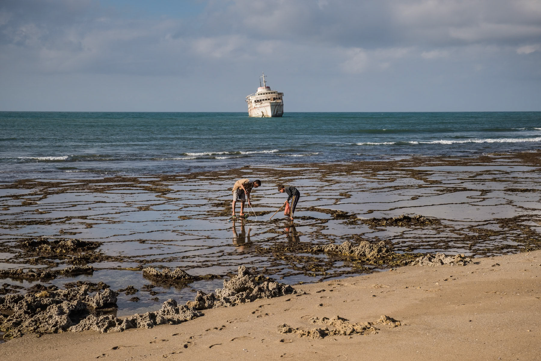 Kids catching shellfish and a stranded ferry that used to sail between the Canary Islands and Tarafaya in Morocco. The wreck has been there since 2008 and the ferry line suspended. Border area between Morocco and the Western Sahara, December 2018