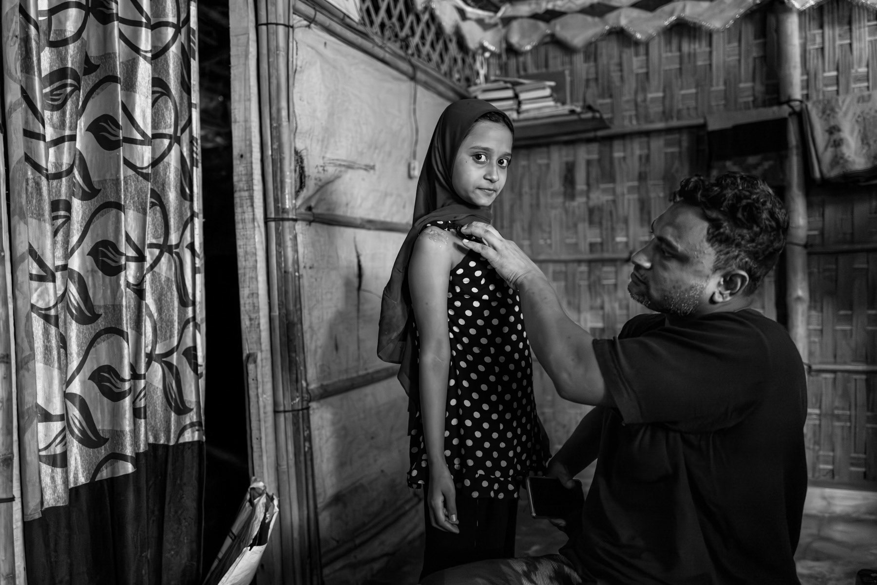 Abu Alom, 36, from Setilla Ward in Maungdaw, stands with his eldest daughter, who shows the healed shrapnel wound on her upper arm from a drone strike carried out by the Arakan Army on 28 July 2024. The attack severely injured him, his wife, and both daughters, forcing the family to flee and reach Bangladesh the same day. Originally from Shabe Bazar—evacuated in 2017—he had resettled in Maungdaw and worked for NGOs before being displaced again by the attack. Kutupalong refugee camp, Ukhiya, Bangladesh, September 2025