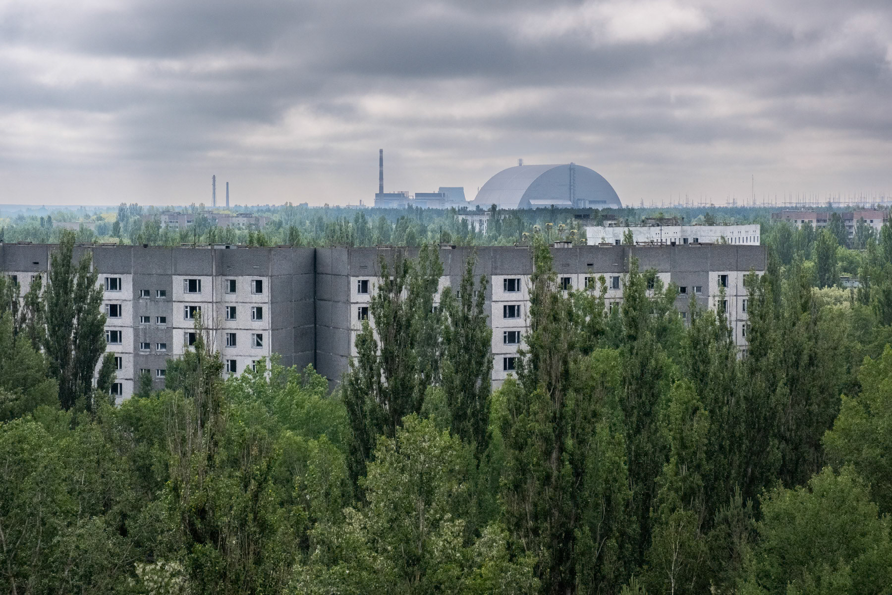 Pripyat residential buildings with reactor number 4 and its protection dome in the background. Pripyat was build as a satelite town only 6 km from the NPP. The complete population of Pipyat was evacuated 36 hours after the disaster to never return again. Pripyat is now an overgrown ghost town where time stood still on 27 April 1986. Pripyat, Chernobyl NPP, Ukraine, June 2021