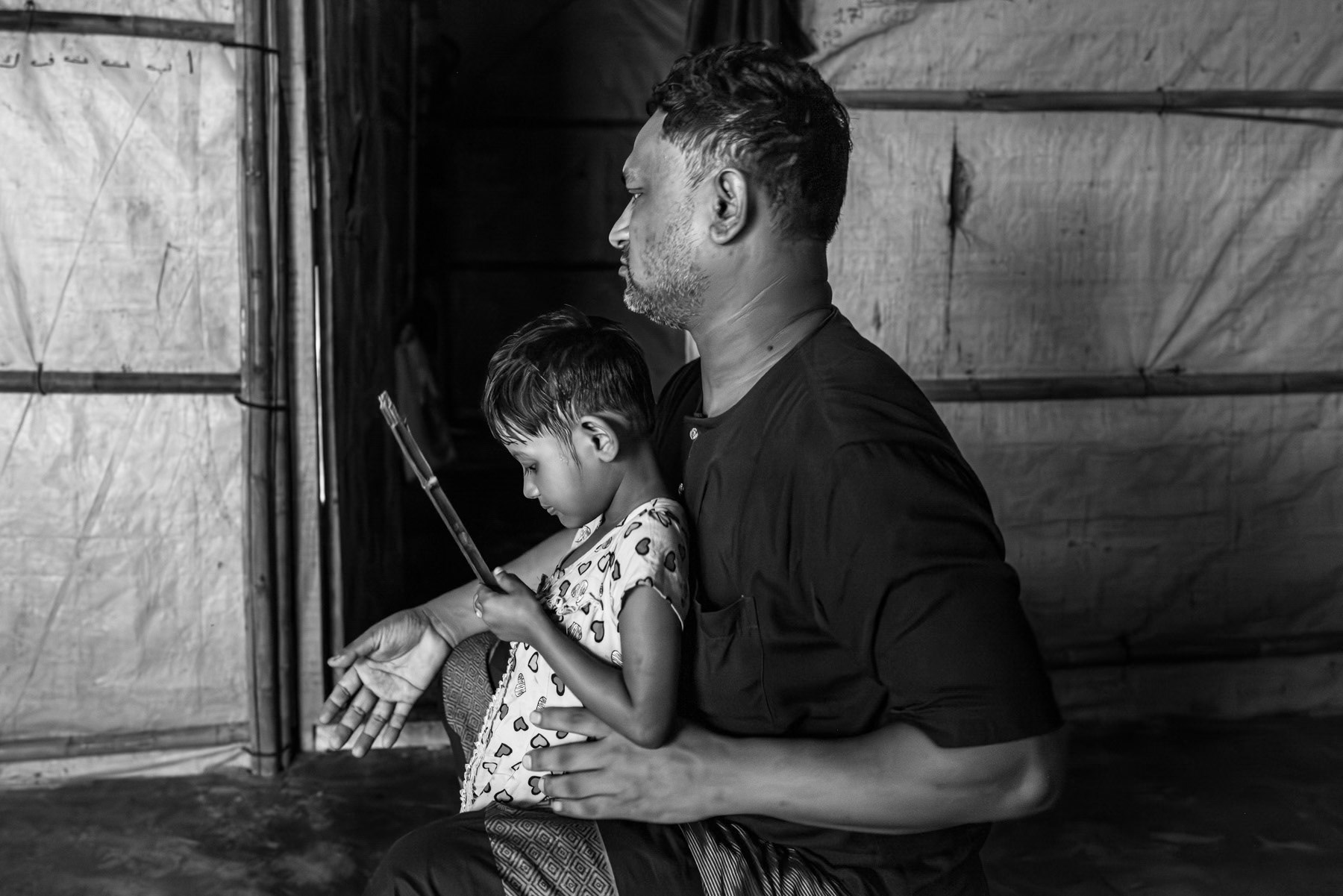 Abu Alom, 36, from Setilla Ward in Maungdaw, stands with his youngest daughter as he recounts how a drone strike carried out by the Arakan Army severely injured him, his wife, and both daughters on 28 July 2024 while they were at home. The family fled immediately and arrived in Bangladesh the same day. Originally from Shabe Bazar—evacuated in 2017—he had resettled in Maungdaw and worked for NGOs before being displaced again by the attack. Kutupalong refugee camp, Ukhiya, Bangladesh, September 2025