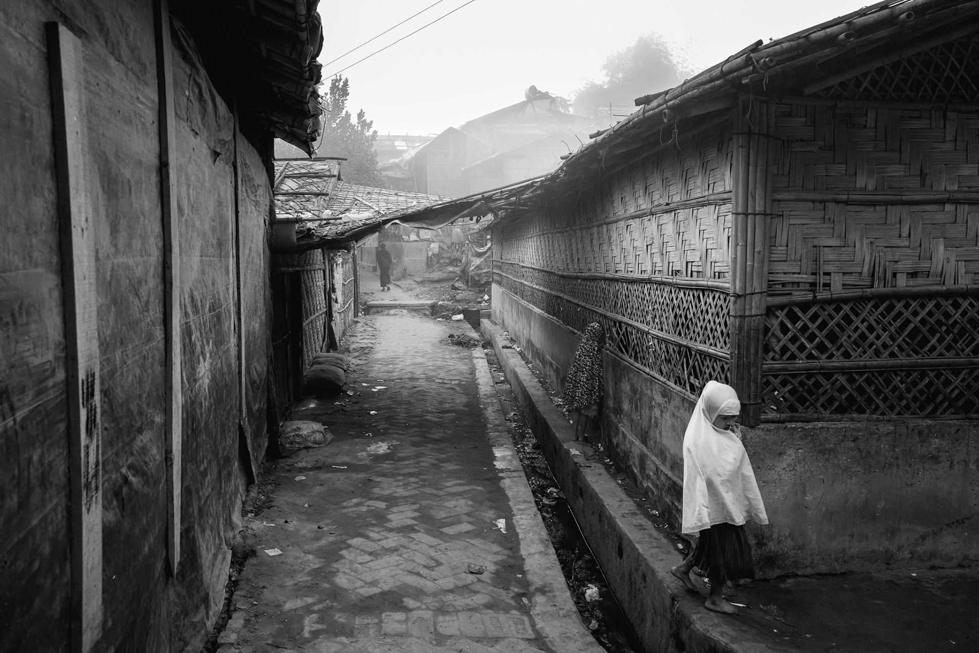 A view of a narrow, brick-paved alleyway between shelters where young girls are walking to a religious school in the early morning. In the camps, religious schools (madrasas) are often the primary form of education available to children. Balukhali camp, Bangladesh, January 2026
