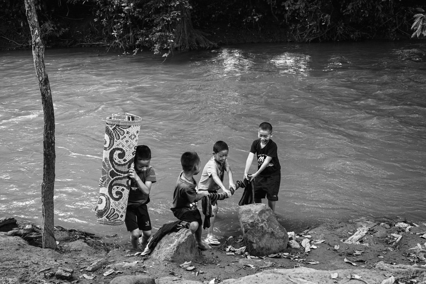 Children washing school mats in a river marking the border with Thailand. This riverside shelter now serves as both home and classroom for displaced Karen children whose villages and schools have been repeatedly bombed by junta forces. Htee Baw Kaw, Karen State, Myanmar, October 2025