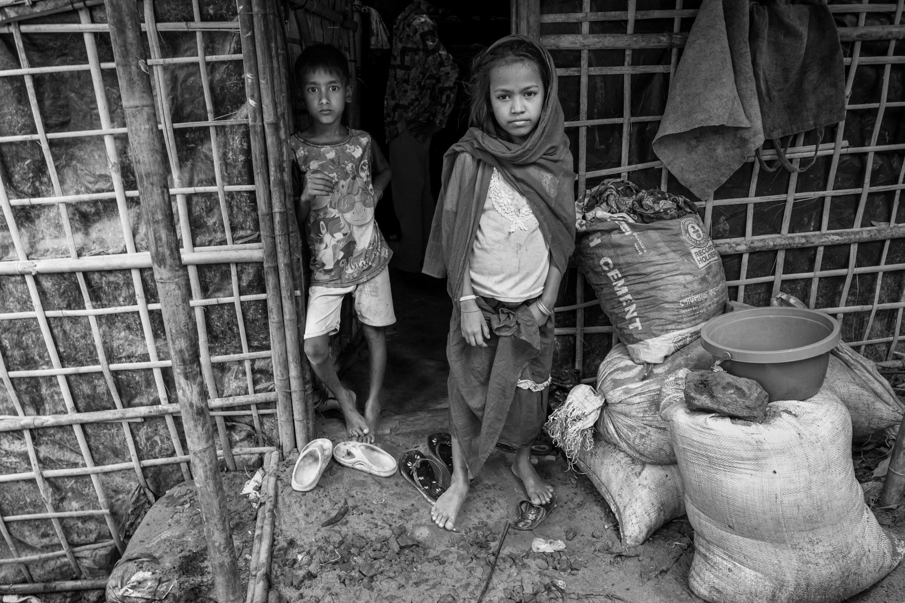 A sister and brother stand in front of their shelter in the camp, part of the more than 150,000 Rohingya who have fled Rakhine since 2024 due to persecution by the Arakan Army. Since 2017, the refugee camps in Bangladesh now host well over 1 million Rohingya, while only a handful remain in Buthidaung and Maungdaw townships. Kutupalong refugee camp, Ukhiya, Bangladesh, September 2025