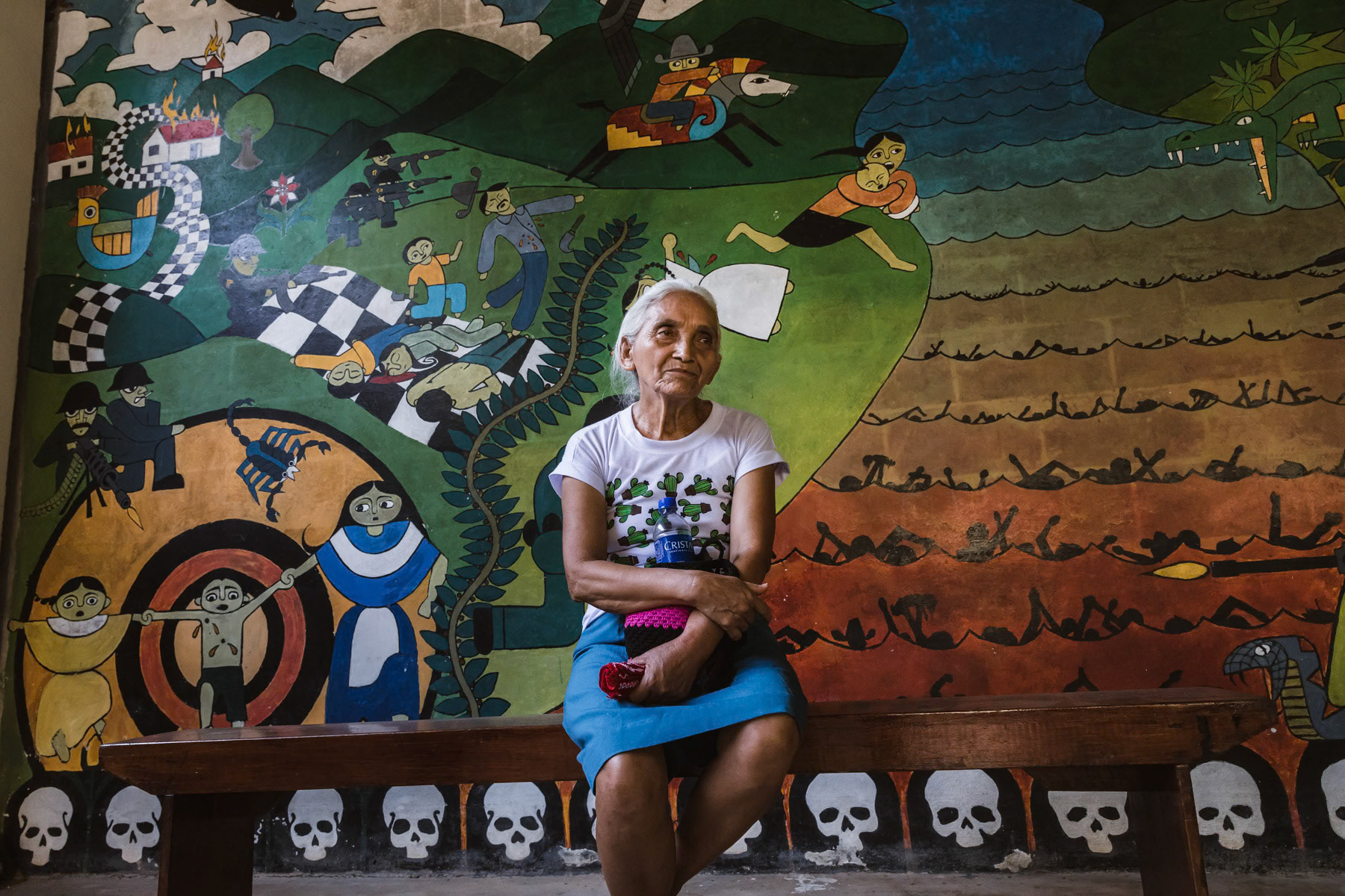 Tomasa Lopez in front of a mural depicting a scene of the Sumpul river massacre. Tomasa from Arcatao witnessed when in 1980 the town and surrounding villages were attacked by government military forces and villagers including children would be randomly executed and hung from trees. Her mother and sister were killed, she fled while being pregnant with her 2 nieces and 2 nephews to the mountains and joined the resistence Farabundo Marti National Liberation Front (FMLN). In 1982 her nieces and nephews were abducted by the military and she lost all track. They were eventually tracked by Pro Busquera, an organisation searching relentlessly for dissapeared children during the civil war, in Spain, France and the US in 1996. They had been sold and adopted through a well established system during the war operated by government military forces and groups in the US and Europe. They have since been reunited at several occasions but not yet in El Salvador. Arcatao, Chalatenango, El Salvador, April 2019