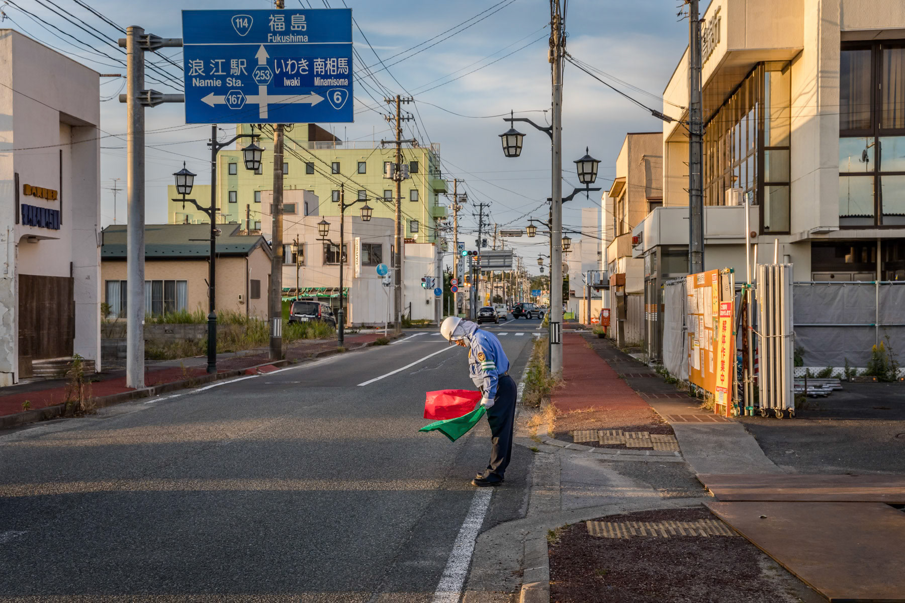 More than eight years after and earthquake, tsunami and one of the most severe nuclear accidents in history, the Japanese prefecture prefecture of Fukushima is trying to get slowly back to normal life where possible. Government officials claim that most areas are safe to return, that radiation levels are equivalent to major world cities and that fresh vegetables and fish are fine to eat. However, the authorities' main problem is to rebuild trust among the local population after being accused of so many coverups and denials about the exact magnitude of the disaster, during and in the aftermath of the nuclear meltdown and explosion. Whereas the earthquake and following tsunami in March 2011 killed 16,000 people. The subsequent nuclear reactor explosion sent radioactive dust over a wide area around the Daichi nuclear power plant, causing more than 160,000 people to abandon and evacuate their homes accross 10% of the Fukushima perfecture. All live, agriculture, fishing and social economical activity ceased in those territories, and many towns and villages became ghost towns in no-go areas. Shortly after the disaster, a massive decontamination effort was started and is presently still under way. It is estimated that several hundreds years will be needed to decommission and decontaminate the Daichi nuclear power plant site itself. Since the disaster most evacuees have gone back to their homes, except for an area of about 3% of the prefecture which remains officially off limits in surrounding forests and towns nearest to the nuclear plant. Okuma and Namie/Futaba, two towns with respectively 11,500 and 21,000 pre-disaster population, and where the nuclear plant is located, have in 2018/2019 been re-opened for people to return living. Only a fraction of the pre-disaster population has returned, few hundreds in Okuma and less than 1,000 in Namie. Workers continue relentlessly to remove and replace contaminated topsoil and other radioactive waste from decomissioned houses and buildings, so that evacuees can return and rebuild their lives. Will they return after having left the area almost 10 years ago is very doubtful, and with almost no economical activity it is impossible for the younger generation to find work. A traffic agent indicating when heavy trucks loaded with construction waste enter the road. Most buildings have been declared unsafe for living and are being brought down. The potentially contaminated building materials are being packed and driven outside the town. Namie, Fukushima Prefecture, Japan, October 2019