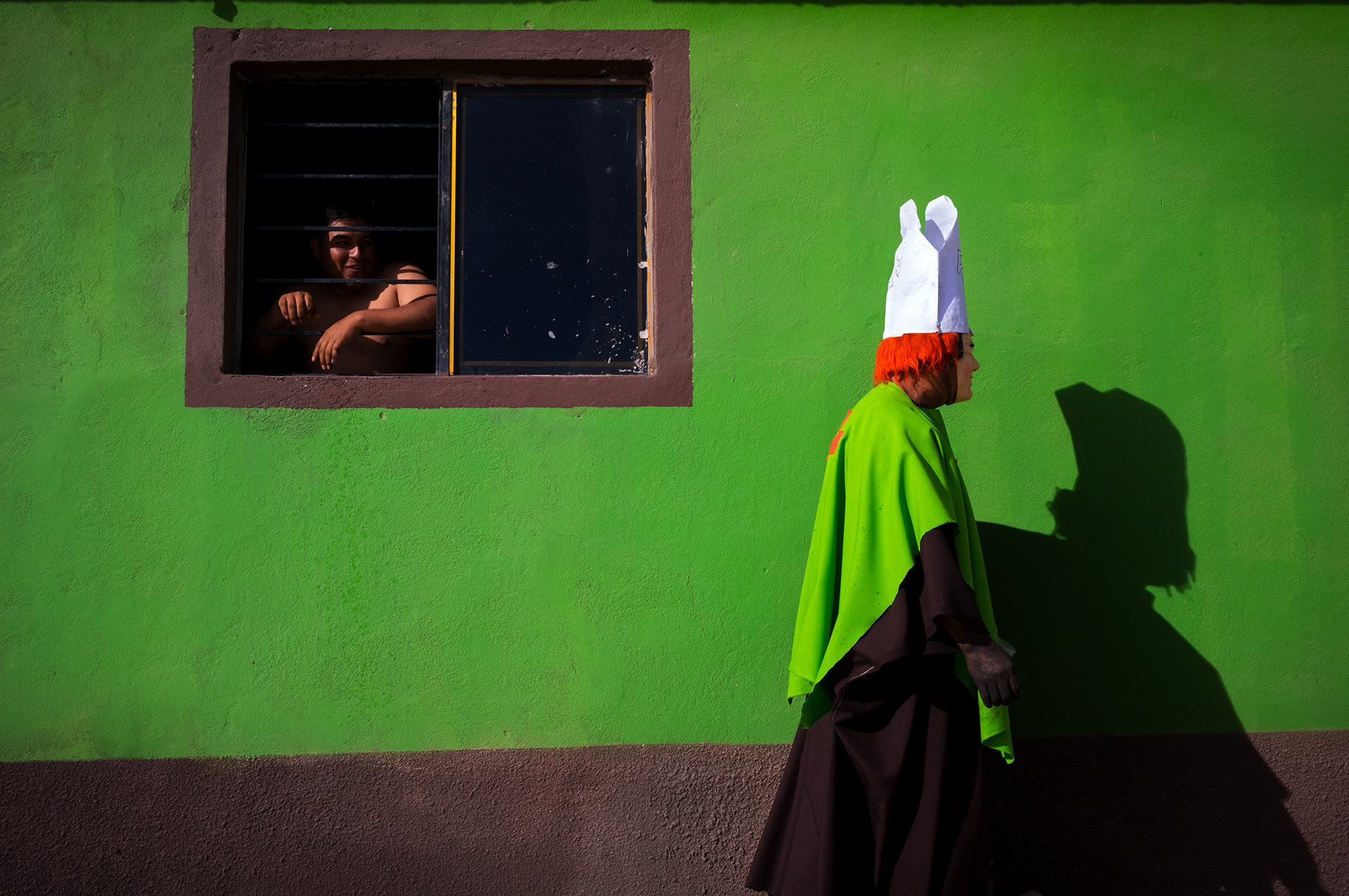 In the Oaxaca community of Santo Tomas Jalieza reveler wearing a green, home made priest costume strides along the bright green wall of a home – just past  the window as the homeowner cautiously peeks out of his window.