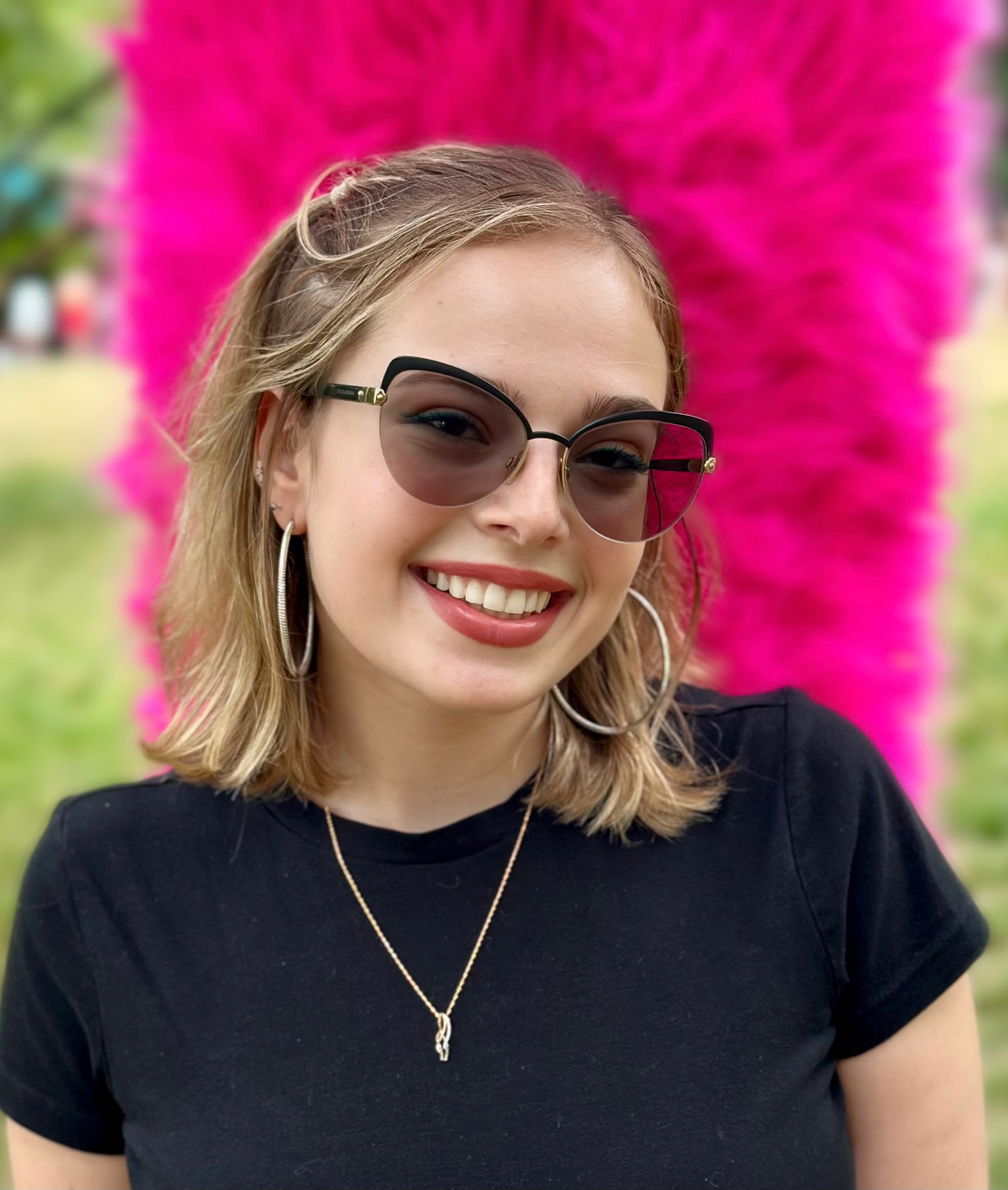 A photo of Sarah wearing a black shirt, glasses, and hoop earrings. She is standing in front of a pink background in a park.