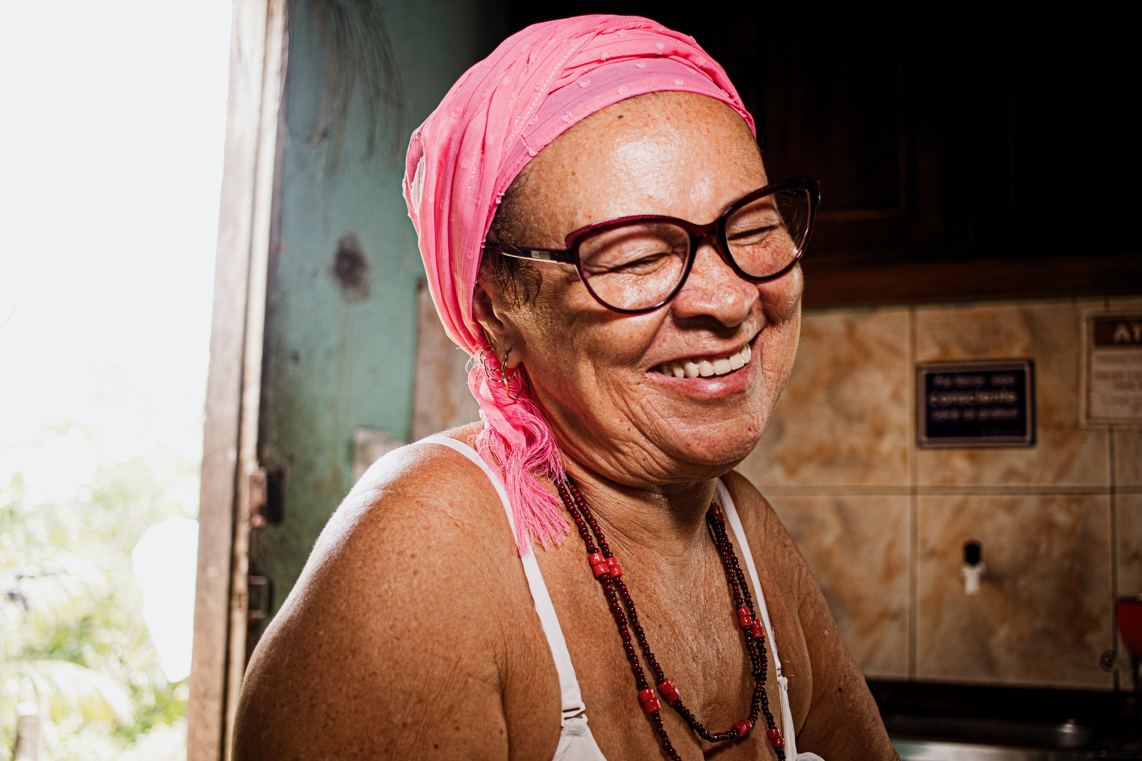 #PraTodosVerem: A fotografia colorida mostra uma imagem mais aproximada de uma senhora branca sorrindo com seus olhos fechados que acompanham seu sorriso. Ela está usando um pano rosa na cabeça, usa óculos de grau e um fio de contas marrom e vermelho no pescoço. A imagem enquadra da altura do busto para cima e mostra as alças brancas da roupa que ela está vestida. Ao fundo, do lado esquerdo, a claridade de uma porta aberta e atrás dela na parte direita da fotografia, uma pia com a parede de cerâmicas em tons claros de marrom, avisos colados nela e abaixo uma torneira preta e branca e parte da cuia da pia é mostrada também. Acima da pia, aparentemente um armário de madeira.