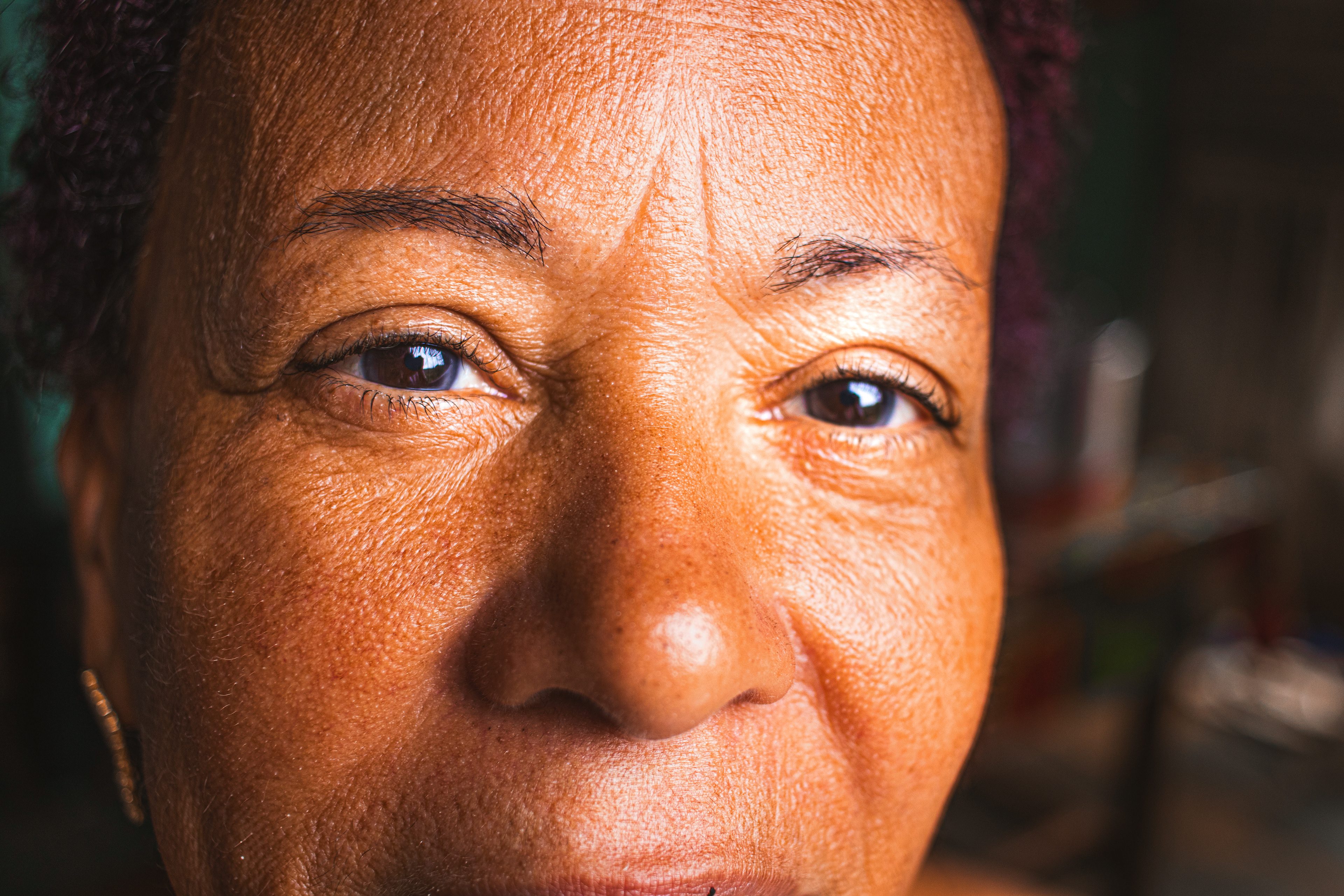 #PraTodosVerem: A fotografia colorida mostra uma mulher negra no centro da fotografia. Ela está encurvada com os braços em direção ao chão de cerâmica branca para pegar as bandeirinhas brancas que enfeitarão o teto do barracão. Ela está descalça, com um vestido longo “tomara que caia”, com estampa de mandalas coloridas. Seu cabelo castanho escuro cacheado está preso para cima e sua expressão é de concentração. No fundo da imagem mostra-se fileiras prontas de bandeirinhas brancas penduradas na altura média do barracão, duas pessoas desfocadas na fotografia, ao fundo uma parede com tom de azul bem claro, um comongol nessa parede e ao lado esquerdo da imagem, ao fundo, três cadeiras brancas empilhadas.