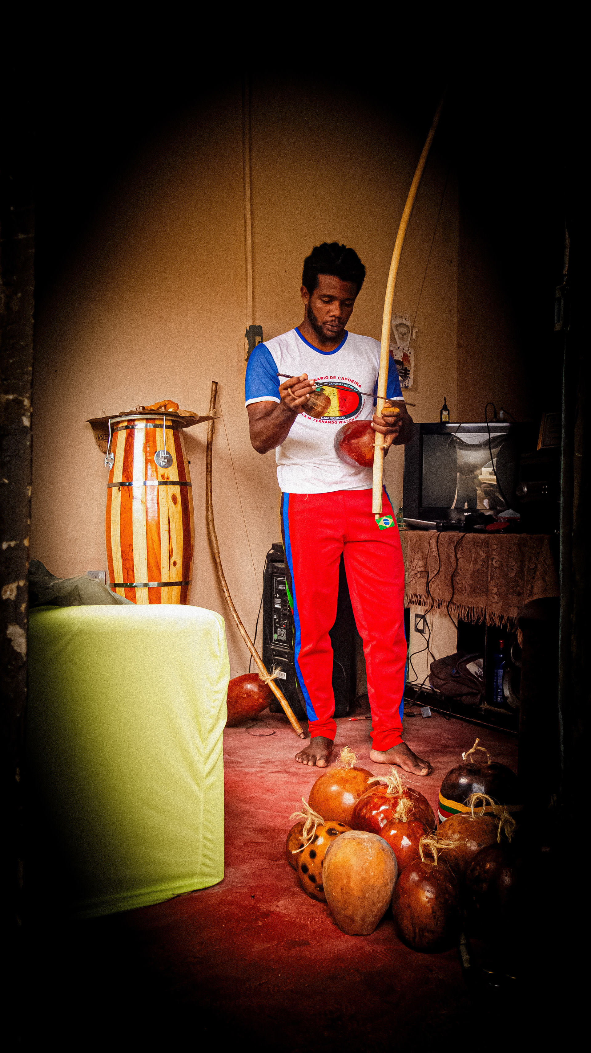 #PraTodosVerem: A fotografia colorida mostra um homem negro com barba e cabelos pretos. Ele está na sala de estar tocando berimbau e na mão direita segura também um caxixi. Está vestido em uma camisa branca com mangas e gola azul, uma camisa com a estampa do símbolo de um grupo de capoeira, calça vermelha com listras verde, preta e azul na lateral. Na perna esquerda a calça tem uma estampa pequena da bandeira do Brasil. Ele está descalço e o chão é vermelho. Na sua frente, no chão da sala, dez cabaças confeccionadas para berimbaus. Na parte esquerda da fotografia mostra a lateral de um sofá com capa verde e do mesmo lado atrás do homem tem um atabaque e um berimbau encostado na parede. Atrás dele uma caixa de som preta e do lado direito da imagem uma mesa forrada com a televisão em cima, controles de videogame e alguns fios conectados. Sob a mesa, uma bolsa ou mochila cinza e uma garrafa azul com tampa vermelha.