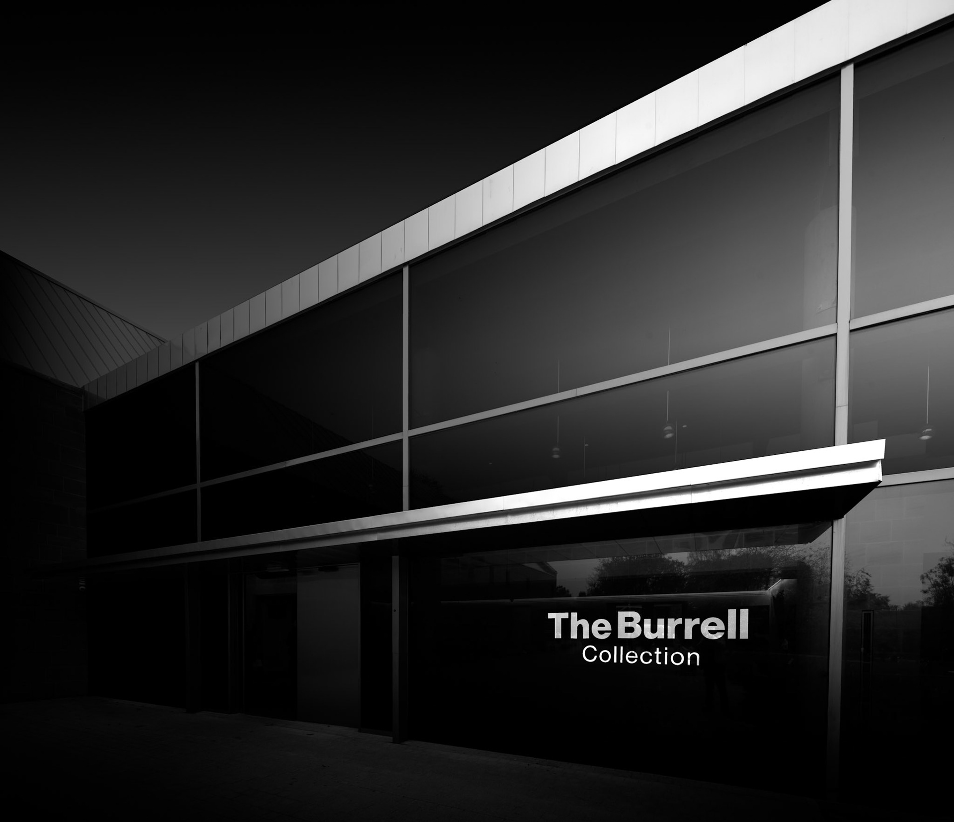 The entrance to the Burrell Collection is reduced here to a carefully structured arrangement of horizontal and vertical lines. The illuminated museum sign provides a small but important point of identification within an otherwise highly abstract architectural composition. Above it, the long white edge of the roof cuts diagonally across the frame, while slender structural columns divide the dark glazed façade into a series of geometric panels. Reflections within the glass further complicate these divisions, allowing the boundary between the building and its surroundings to become uncertain. Light has been deliberately restricted to a handful of architectural elements, drawing attention to the precision and rhythm of the structure rather than revealing the whole façade. The resulting image sits somewhere between architectural record and abstraction: recognisably the entrance to the Burrell, but primarily concerned with line, proportion, reflection and the relationship between illuminated surfaces and deep shadow.