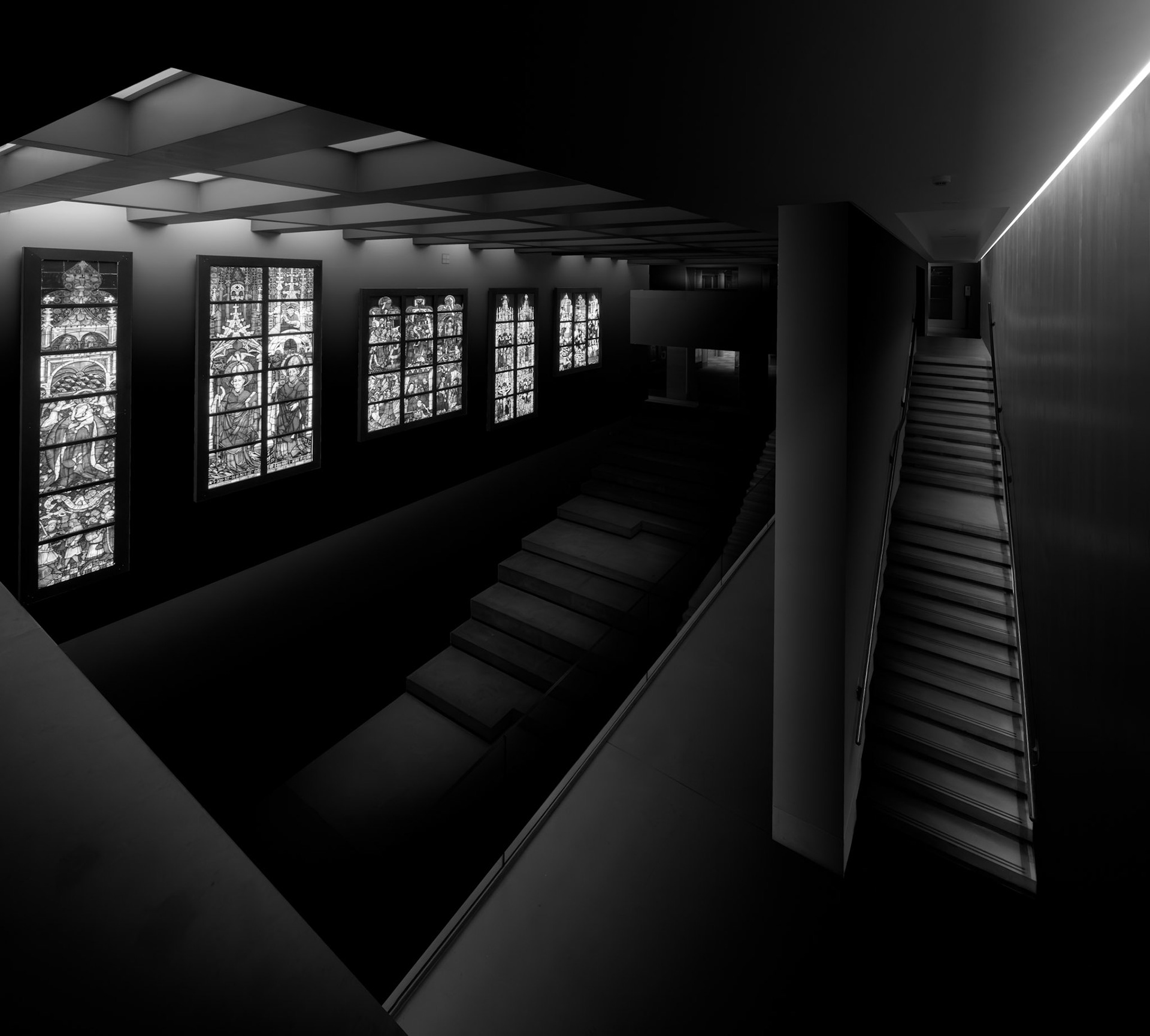 This elevated view reveals the complex relationship between galleries, circulation and display within the refurbished Burrell Collection. A staircase descends through the right side of the composition while a second flight rises beyond it, creating opposing diagonal movements through the otherwise strongly rectilinear architecture. To the left, a sequence of illuminated display panels provides the principal source of light, repeating across the dark gallery like a series of windows. The viewpoint allows several levels of the museum to be understood simultaneously, but much of the architecture remains deliberately concealed in shadow. This selective illumination turns a potentially complicated interior into a carefully ordered arrangement of planes, lines and luminous rectangles. The photograph is concerned less with individual exhibits than with the architecture through which they are encountered. In monochrome, the museum becomes a layered spatial composition in which light describes both the collection and the routes visitors take around it.