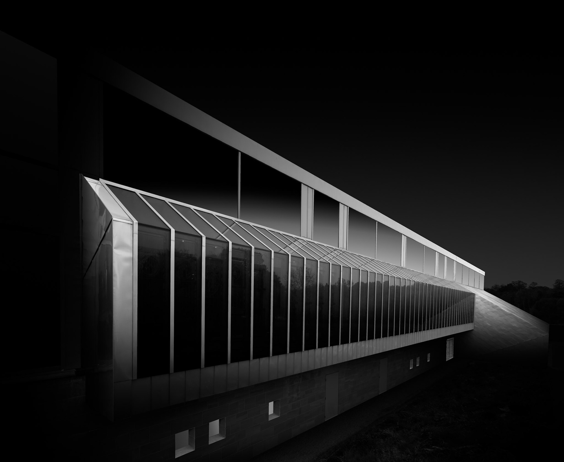 The long glazed elevation of the Burrell Collection is viewed obliquely, turning its repeating structural framework into the dominant element of the photograph. A sequence of illuminated vertical fins recedes towards the far end of the building, their diminishing spacing creating a powerful sense of perspective and movement. Above them, the roofline sweeps diagonally across the composition, while the darker structural frame behind provides a restrained counterpoint to the brighter façade. Very little of the surrounding landscape is allowed to intrude, leaving the building isolated against an almost completely black background. The result emphasises one of the Burrell's defining architectural contrasts: the lightness and transparency of its extensive glazing set against the more substantial elements of the museum. Through the monochrome treatment and selective use of light, a functional elevation becomes an abstract sequence of luminous lines, with repetition and perspective giving the architecture an almost rhythmic quality.