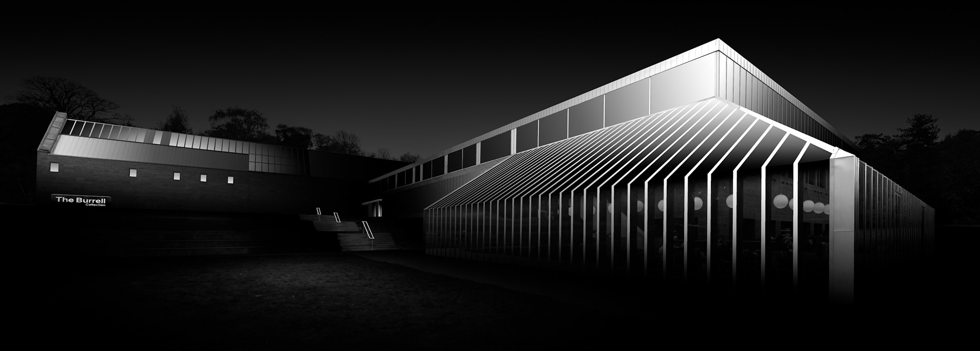 The Burrell Collection stretches across the landscape in this panoramic exterior view, bringing together two contrasting aspects of its architecture. To the left, the substantial masonry volume of the museum sits quietly in darkness, punctuated by a small sequence of illuminated openings. To the right, the glazed gallery projects forward as a much lighter and more transparent structure, its repeating vertical framework picked out dramatically against the night. The brightest light is concentrated around the roofline and upper corner, from where it appears to travel down through the rhythm of the façade. The wide format emphasises the dialogue between these two architectural forms: solid and transparent, heavy and light, enclosed and open. Surrounding detail has been deliberately suppressed so that the building appears almost detached from its setting. What remains is an architectural study constructed from repetition, proportion and carefully controlled illumination, with the Burrell emerging gradually from darkness.