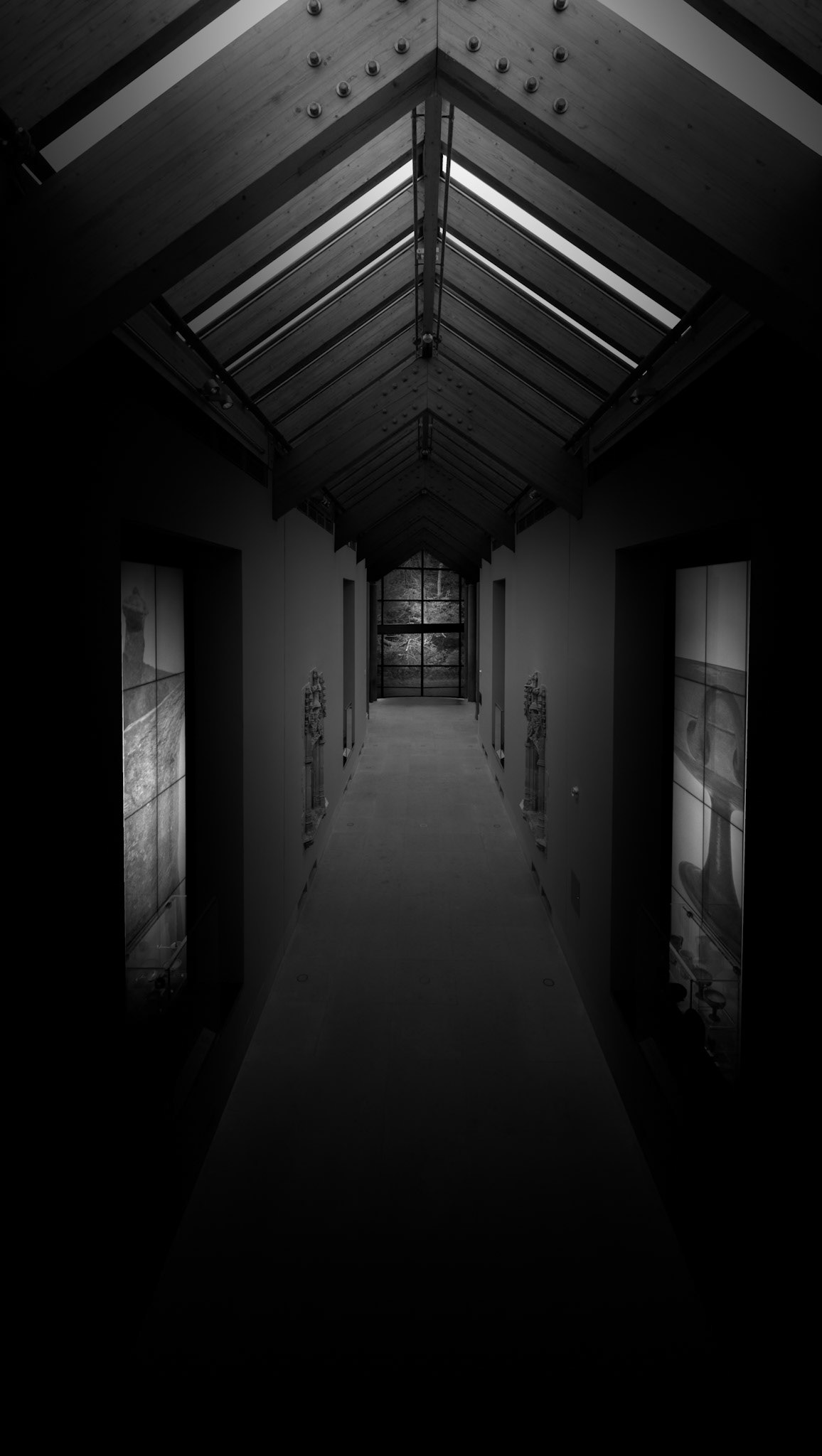 A narrow passage within the Burrell Collection becomes an exercise in perspective, symmetry and controlled light. The steeply pitched glazed roof establishes a strong central axis, its repeating framework drawing the eye towards the far end of the corridor. On either side, display cases and architectural openings emerge only partially from the surrounding darkness, allowing the illuminated roof structure and distant artwork to dominate the composition. The vertical format accentuates the enclosed character of the space and strengthens the sensation of moving forward through it. Rather than presenting the museum as a sequence of brightly illuminated galleries, the photograph concentrates on the quieter spaces connecting them. The restrained black-and-white treatment reduces the architecture to its essential geometry and creates a deliberate tension between what is revealed and what remains hidden. The corridor consequently becomes both a physical route through the Burrell and a visual transition between darkness, light and the collection beyond.
