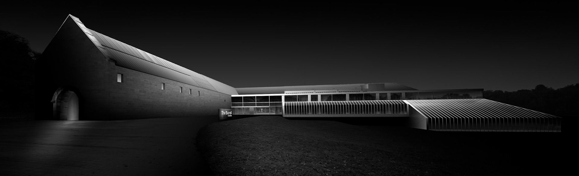 The Burrell Collection sits low against the landscape of Pollok Country Park, its long horizontal form emphasised in this panoramic view. The museum combines substantial areas of masonry with lighter glazed structures, creating an architecture that alternates between solidity and transparency. Here, most of the surrounding landscape has been allowed to recede into darkness, leaving the building defined primarily by carefully controlled areas of light. Illumination catches the sloping roof of the main masonry volume before travelling across the entrance and along the repeating structure of the glazed elevation. The deliberately restrained monochrome treatment removes much of the building's immediate context, transforming it into an arrangement of planes, lines and tonal relationships. Rather than documenting the museum conventionally, the photograph presents the Burrell as a quiet sculptural presence emerging from the surrounding darkness.