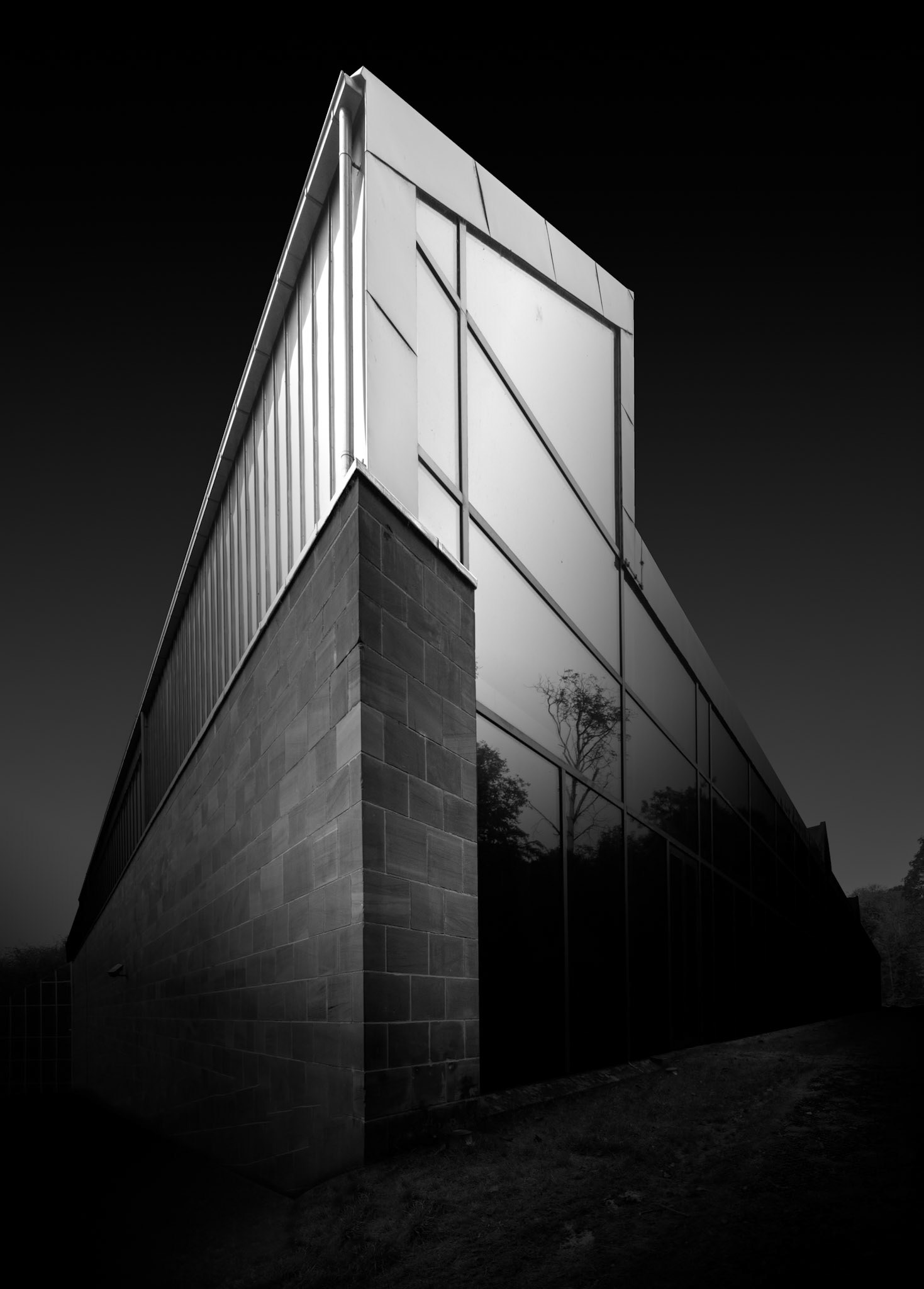 A corner of the Burrell Collection rises sharply through this vertical composition, transforming a relatively small section of the building into a monumental architectural form. The viewpoint is positioned close to the masonry base, allowing the wall to climb through the frame before meeting the much lighter glazed structure above. Strong converging lines exaggerate the height and depth of the building, while the illuminated upper panels appear almost suspended against the dark sky. The contrast between rough, substantial masonry and smooth, luminous glazing encapsulates one of the recurring architectural relationships within the Burrell: solidity meeting transparency. Almost all environmental context has been removed through the deep monochrome treatment, concentrating attention entirely on material, geometry and light. Seen from this unusual angle, the museum becomes difficult to read at a conventional scale, allowing a familiar piece of architecture to operate instead as an abstract composition of planes, edges and sharply defined tonal transitions.