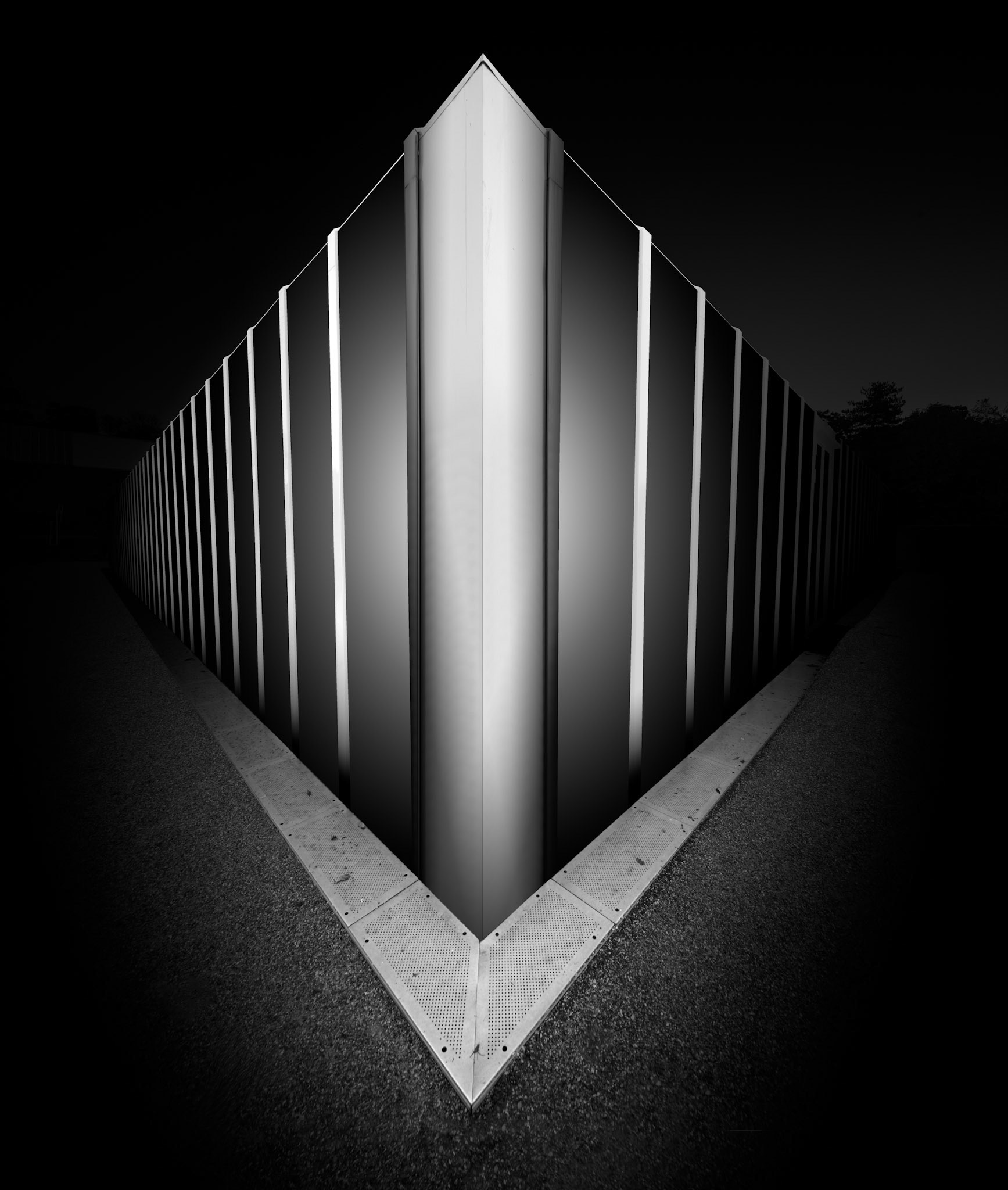 Two glazed elevations of the Burrell Collection converge dramatically towards the centre of this photograph, creating an almost perfectly symmetrical architectural composition. The corner of the building forms a bright vertical blade rising from the foreground, while repeating illuminated fins extend backwards on either side and gradually disappear into darkness. The unusually close viewpoint removes almost every conventional clue to the building's overall form, allowing the façade to become an abstract pattern of light, repetition and perspective. At ground level, textured paving forms a subtle triangular counterpoint to the structure above, reinforcing the central axis and leading directly towards the illuminated corner. The deep black-and-white treatment further separates the building from its surroundings, leaving only those surfaces necessary to describe its geometry. The result is less a conventional representation of the Burrell Collection than a study of architectural rhythm, symmetry and light, reducing the museum to an almost sculptural object.