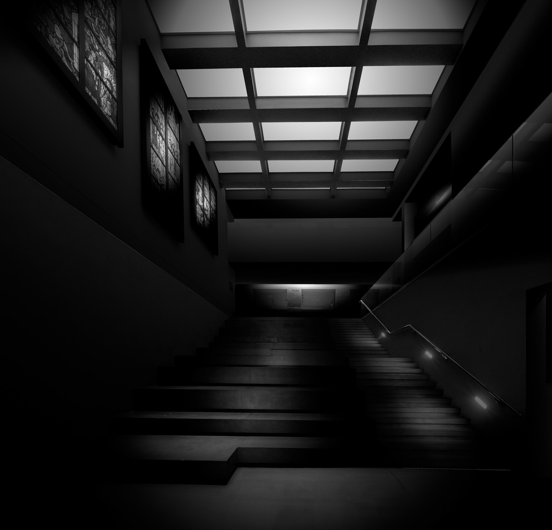 Light descending through the ceiling structure defines this staircase within the Burrell Collection. Rather than attempting to describe the complete interior, the photograph concentrates on the transition between levels, using the illuminated ceiling and subtly lit stair edges to construct a pathway through an otherwise dark space. The strong rectangular geometry overhead contrasts with the diagonal movement of the stairs, while the walls and handrails gradually disappear into deep shadow. A small illuminated area at the centre provides a distant focal point and gives the composition an unexpected sense of depth. The monochrome treatment strips the scene of distraction and emphasises the architectural relationship between light, surface and circulation. What might ordinarily be regarded simply as a functional route through the museum becomes the subject in its own right: an enclosed architectural space in which illumination quietly guides movement and reveals just enough structure for the viewer to understand the route ahead.