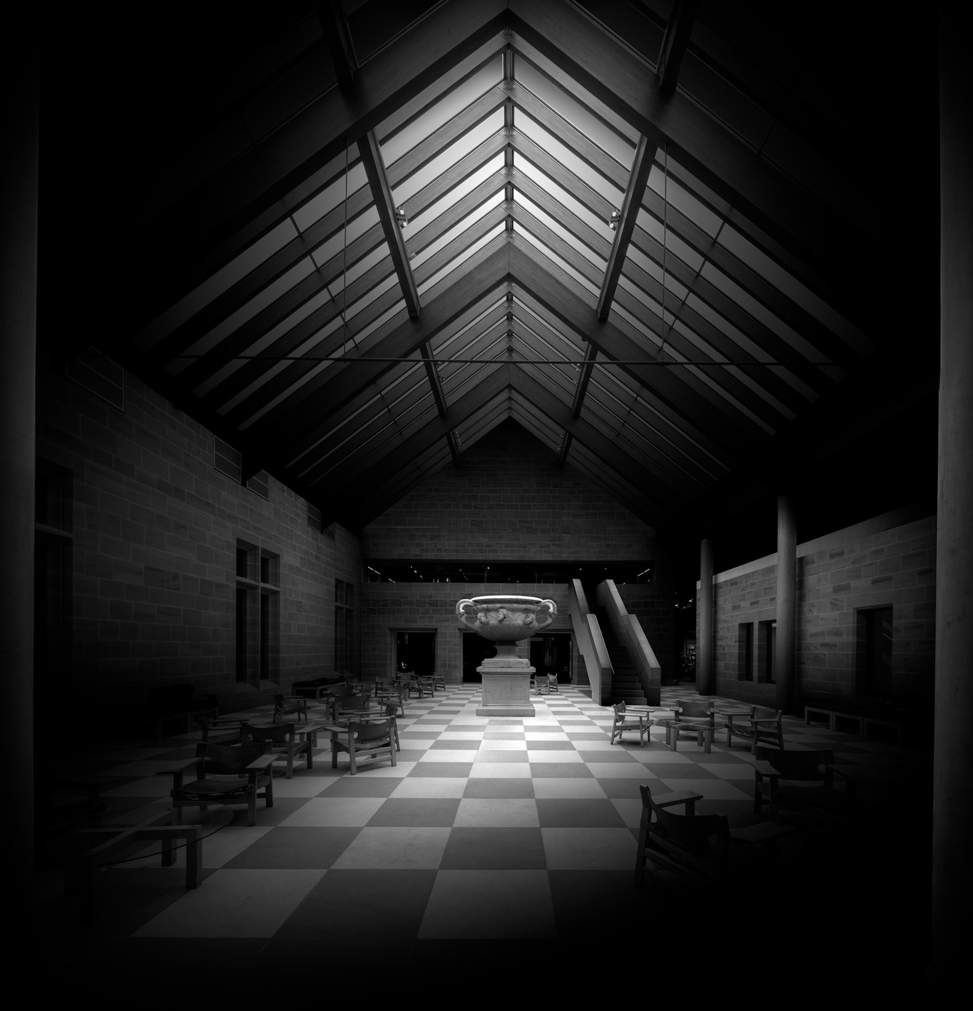 One of the Burrell Collection's distinctive internal spaces is viewed along its central axis, with the architecture itself framing the collection beyond. The pitched glazed roof creates a strong sequence of converging lines that draws the eye deep into the gallery, while the chequered floor reinforces the perspective from below. Heavy masonry walls and the repeating structure overhead establish an almost processional quality, leading towards the illuminated exhibits at the far end of the room. Much of the immediate interior has deliberately been allowed to fall into shadow, concentrating attention on the central route and the objects revealed by light. In monochrome, the relationship between the museum's architecture and its collection becomes particularly pronounced: the building is not simply a neutral container for the exhibits but actively determines how they are encountered. The photograph uses symmetry, darkness and controlled illumination to turn the gallery into a quiet, almost contemplative space.