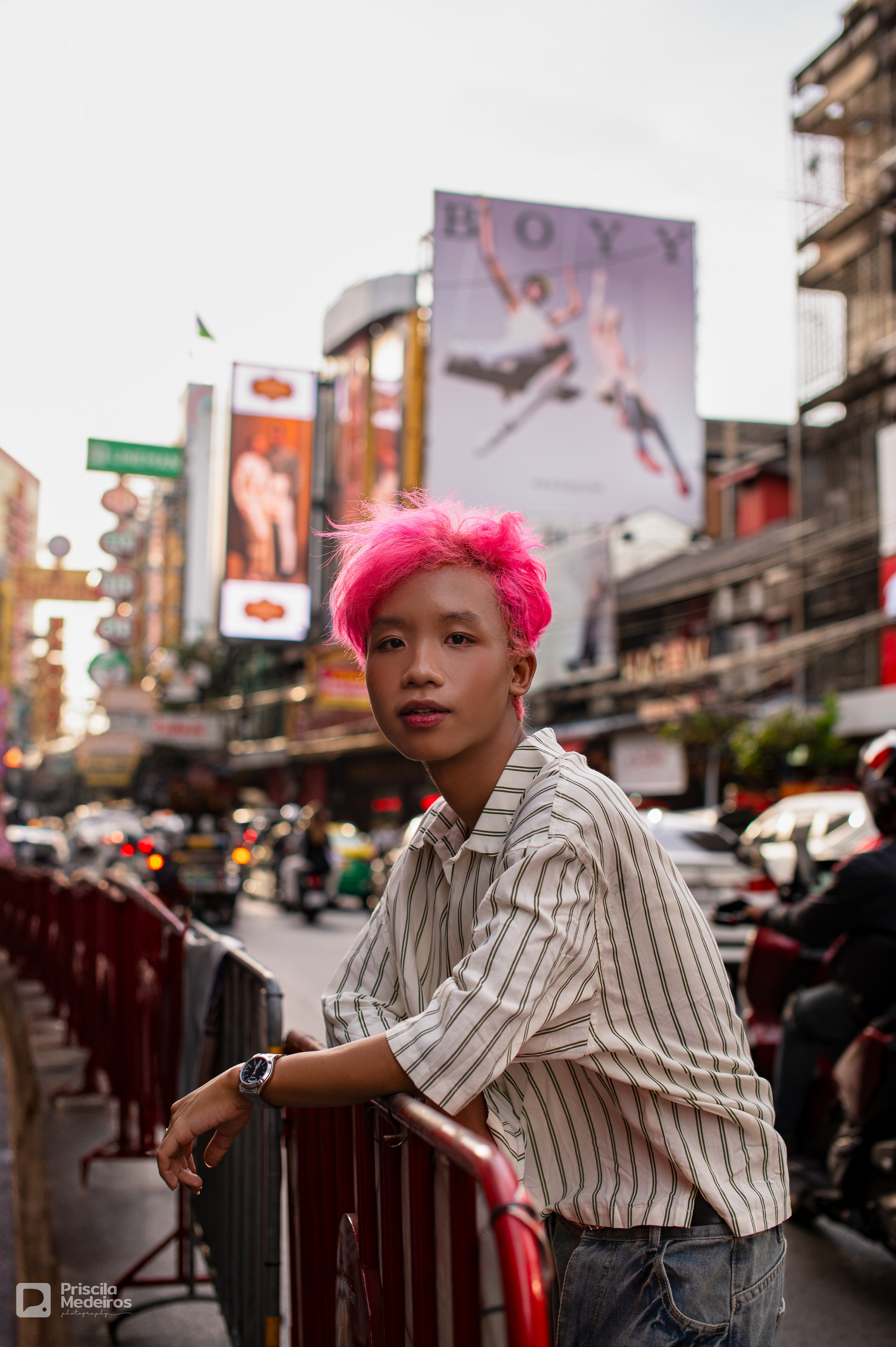 Portrait of a stylish person with pink hair standing on a busy street in Bangkok, Thailand, with blurred city lights and urban billboards in the background.
