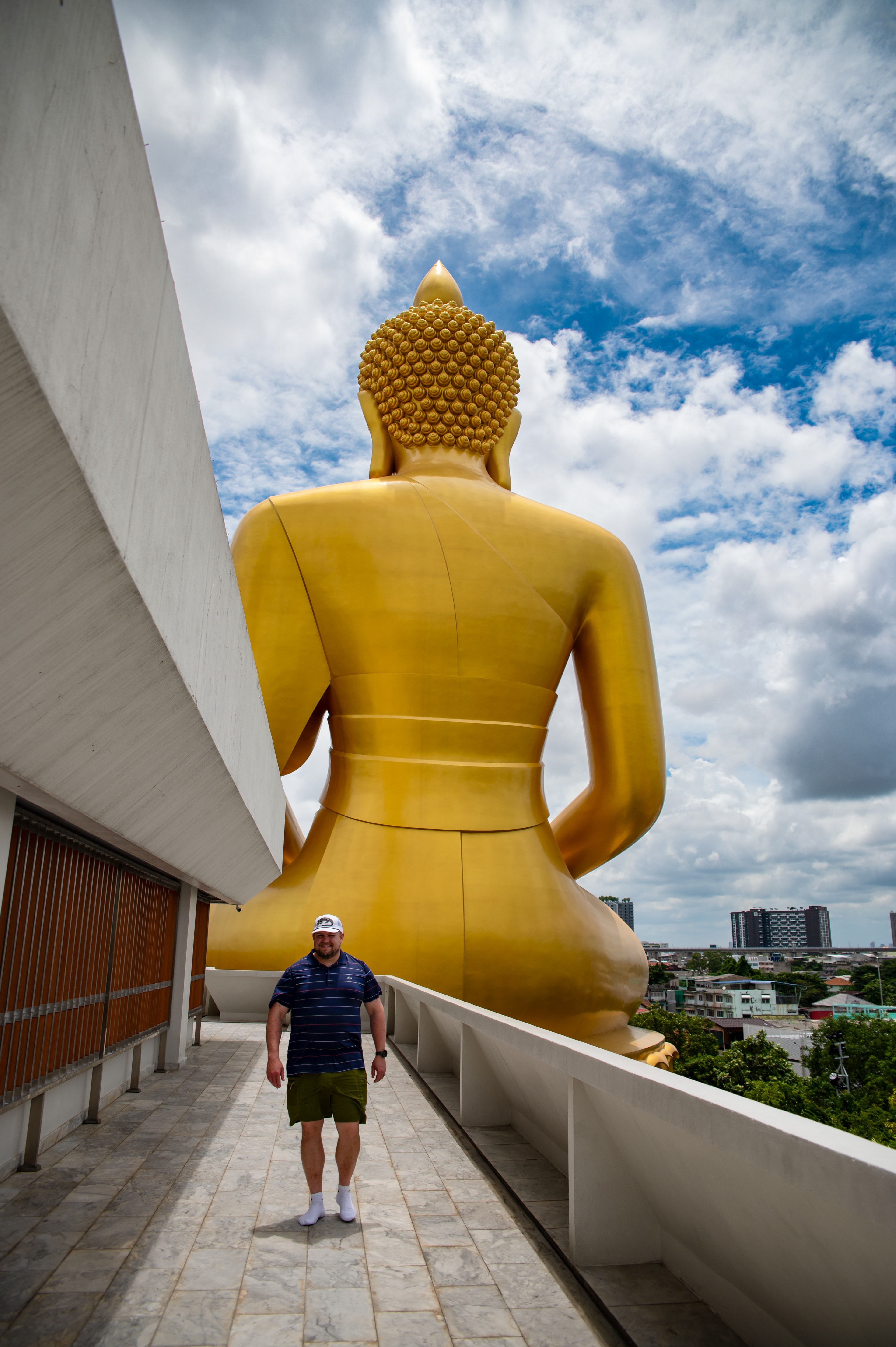 A man walking on a walkway next to a massive golden Buddha statue in Bangkok, Thailand, with a cityscape in the background.
