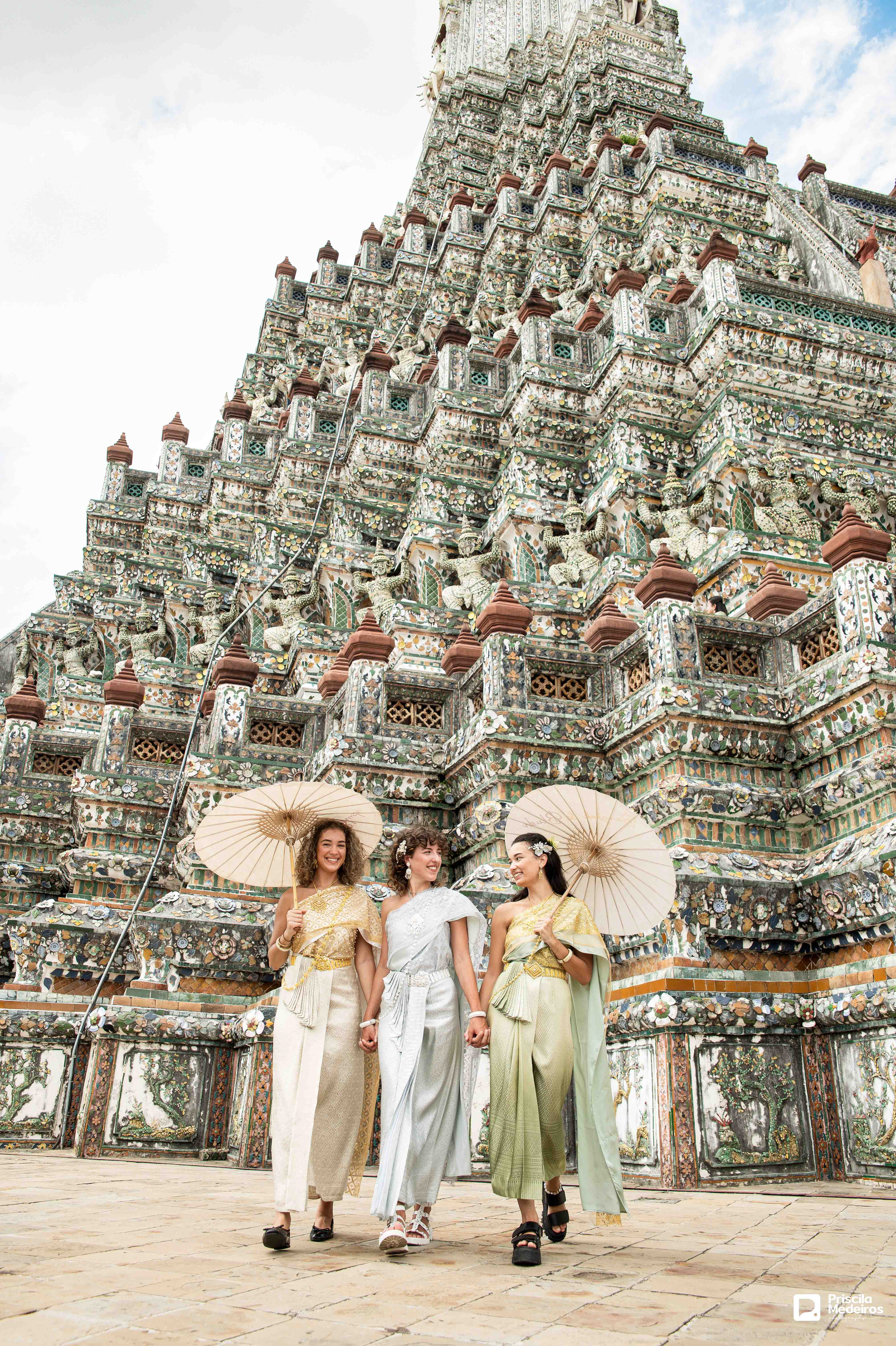 Three women in traditional Thai silk dresses (Chut Thai) holding umbrellas and walking in front of a giant historic landmark in Bangkok; professional group travel photography in Thailand.