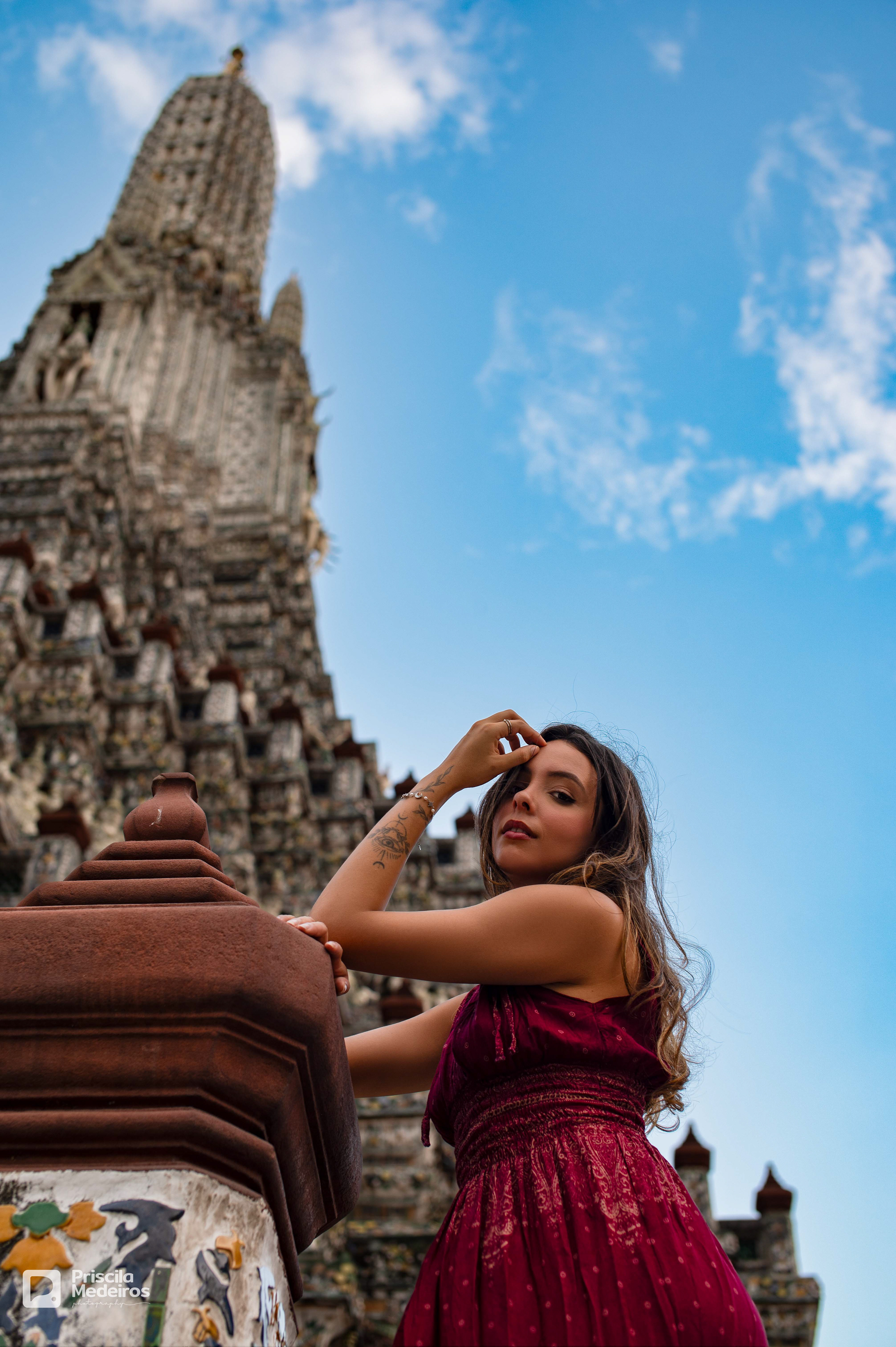 A woman in a dark red dress posing for a professional portrait at Wat Arun Temple in Bangkok, Thailand, with the ornate Khmer-style prang in the background.