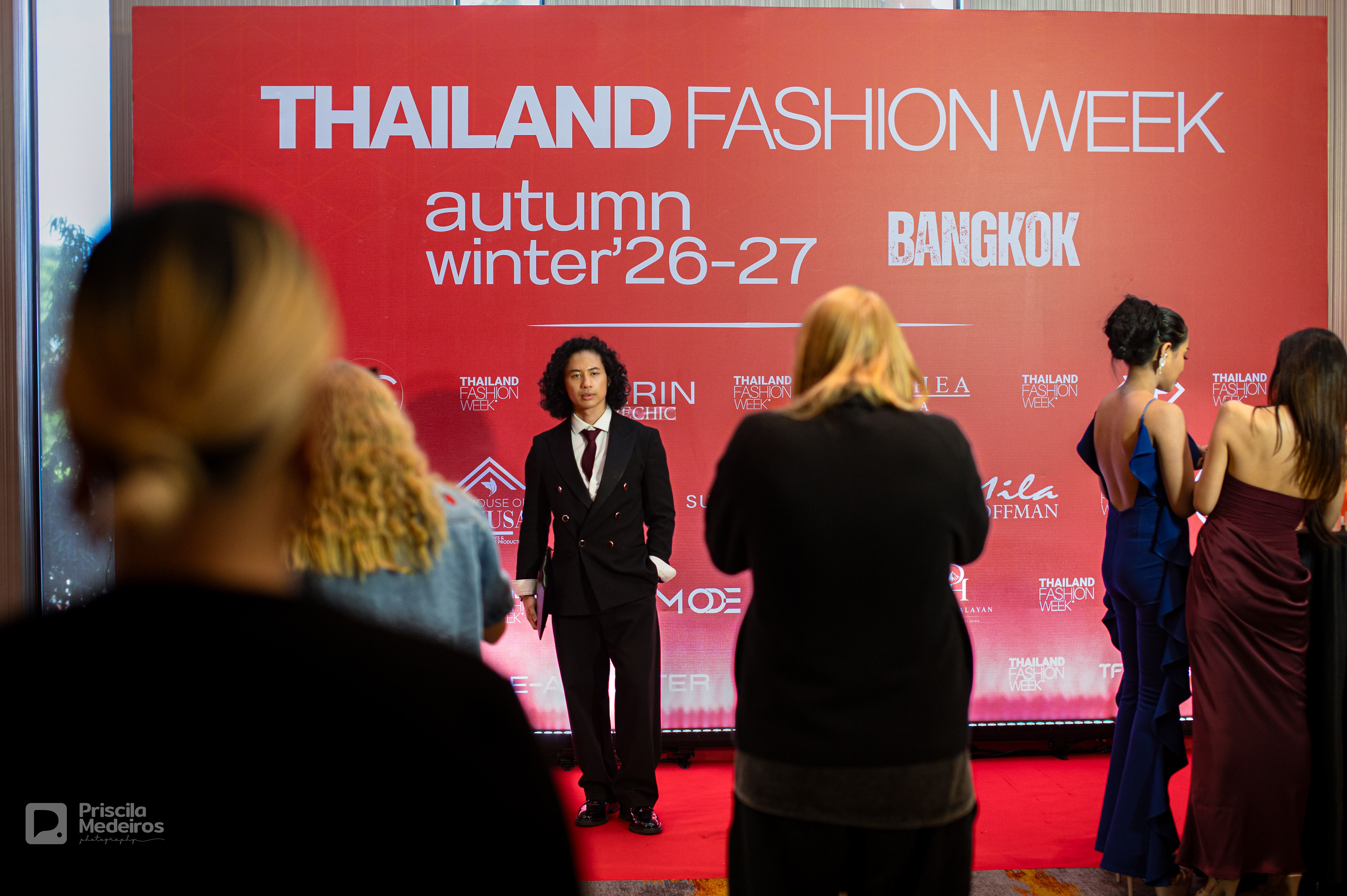 A man posing on the red carpet at the Thailand Fashion Week Autumn Winter 26-27 event in Bangkok, with attendees and event branding in the background.