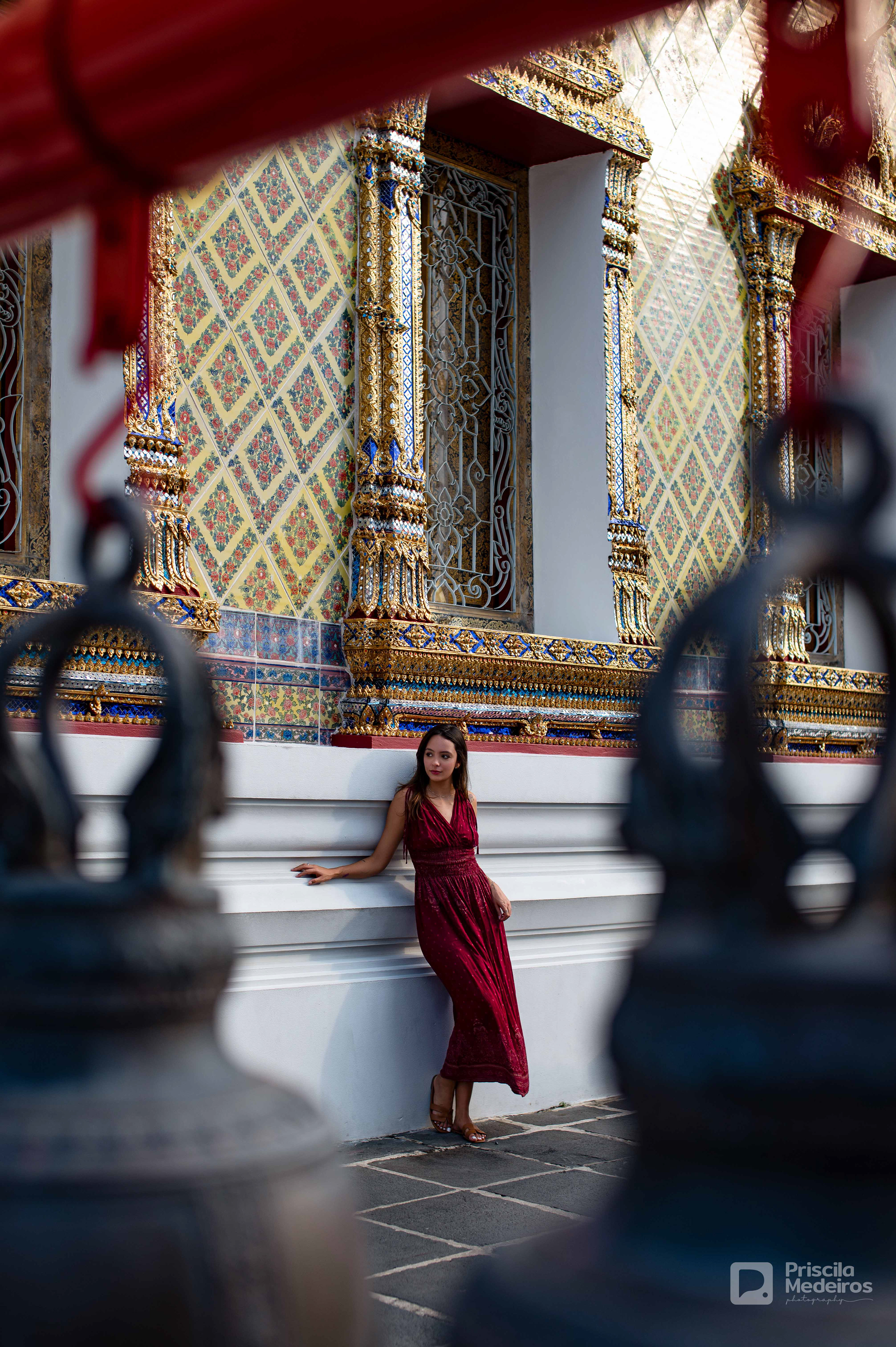 A woman in a red dress standing against an ornate temple wall in Bangkok, captured through a framed foreground of traditional temple bells.
