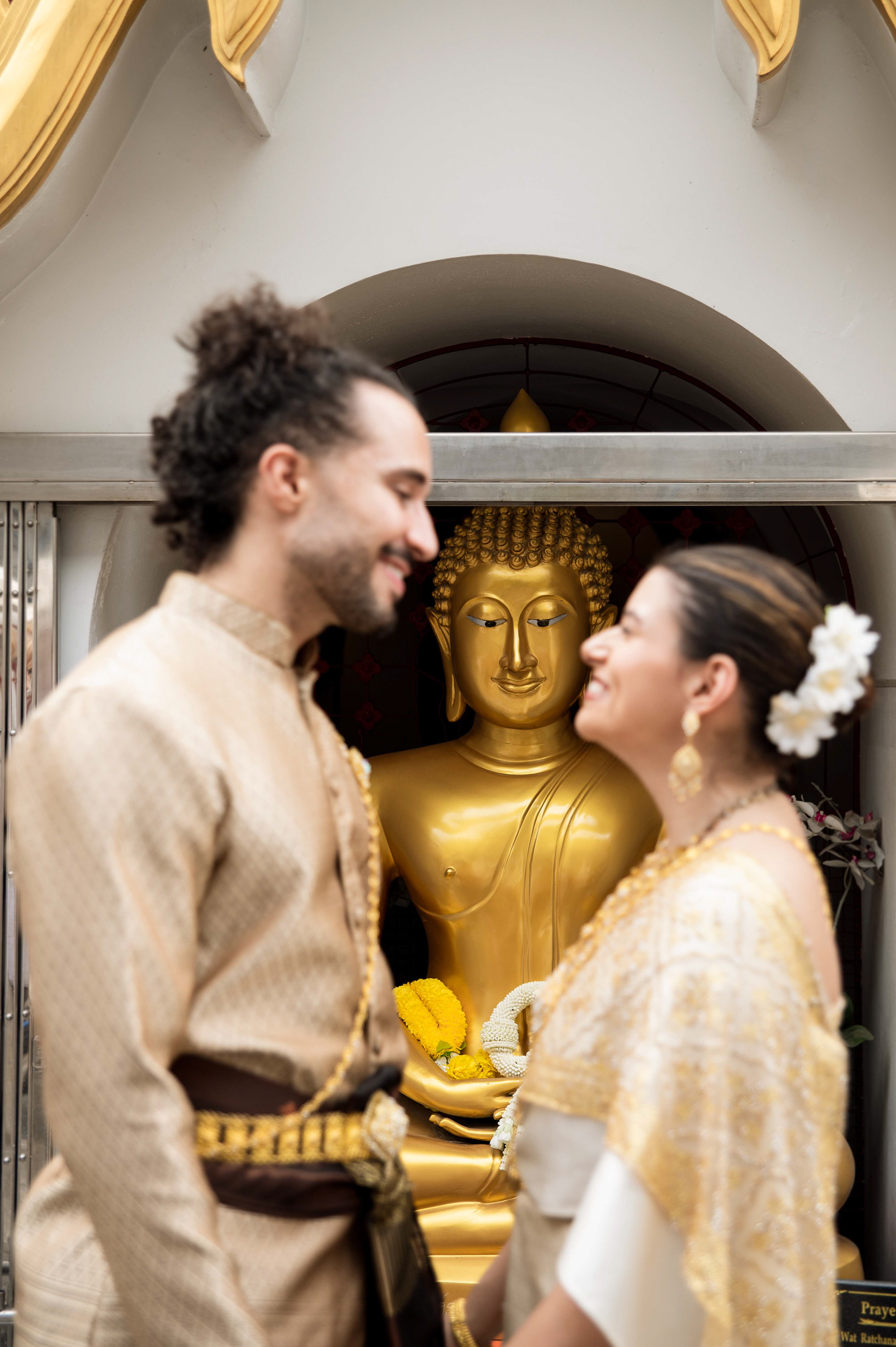 A couple wearing traditional Thai wedding attire smiling at each other in front of a golden Buddha statue at a temple in Bangkok.
