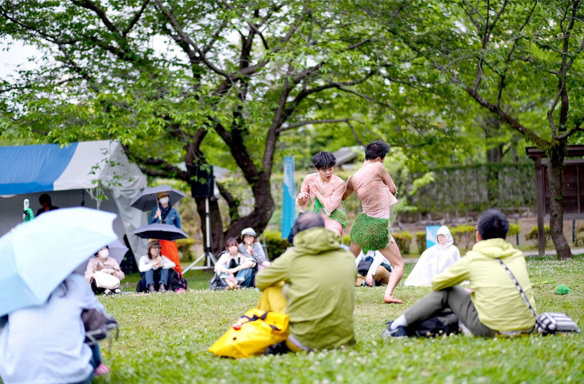芝生の上で踊る二人の男性と、芝生に座りながら鑑賞する人々