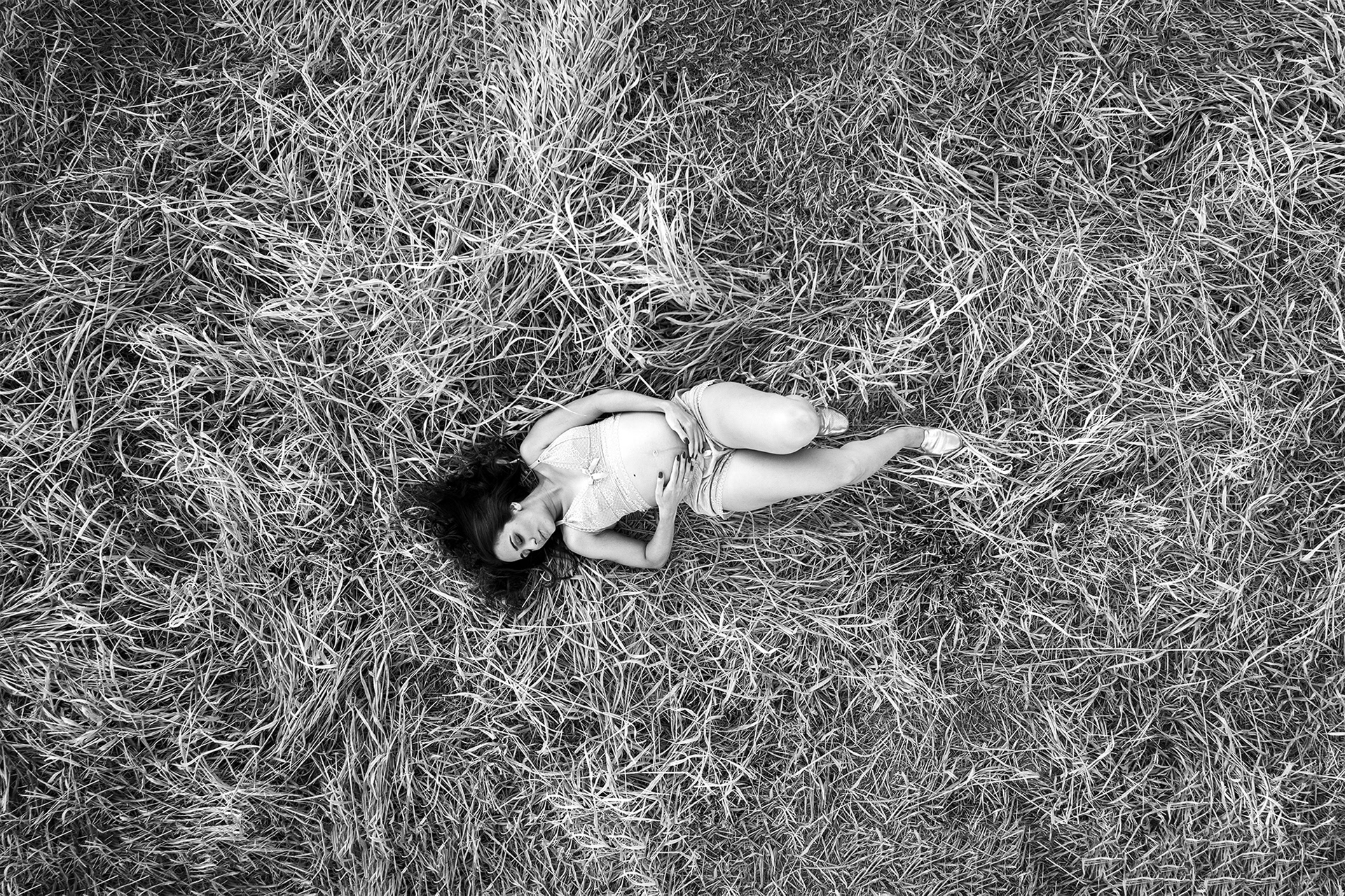 imagem de uma Gestante realizada com um Drone , ela esta deitada no campo e a foto foi tirada de cima com um drone, ela esta com as mãos na barriga bem ao centro da foto