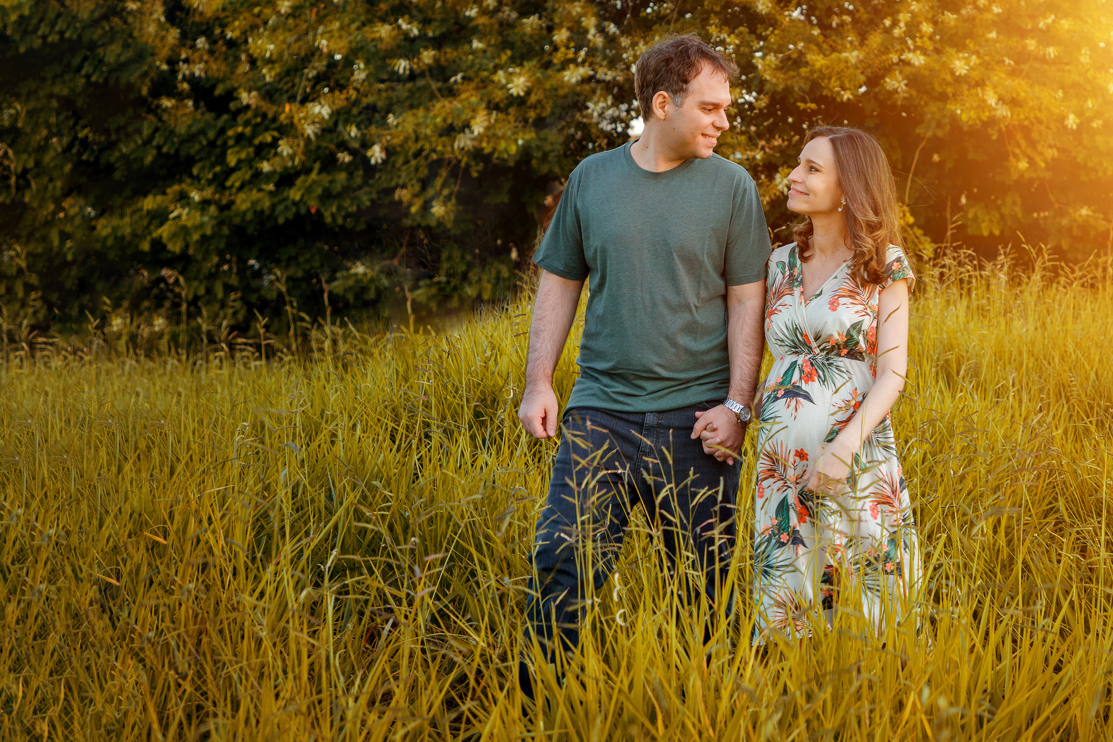 um casal onde ela esta gravida andando de mãos dadas em meio ao um campo gramado , estão trocando olhares e caminhando a luz do sol esta bem amarela devido ao por do sol
