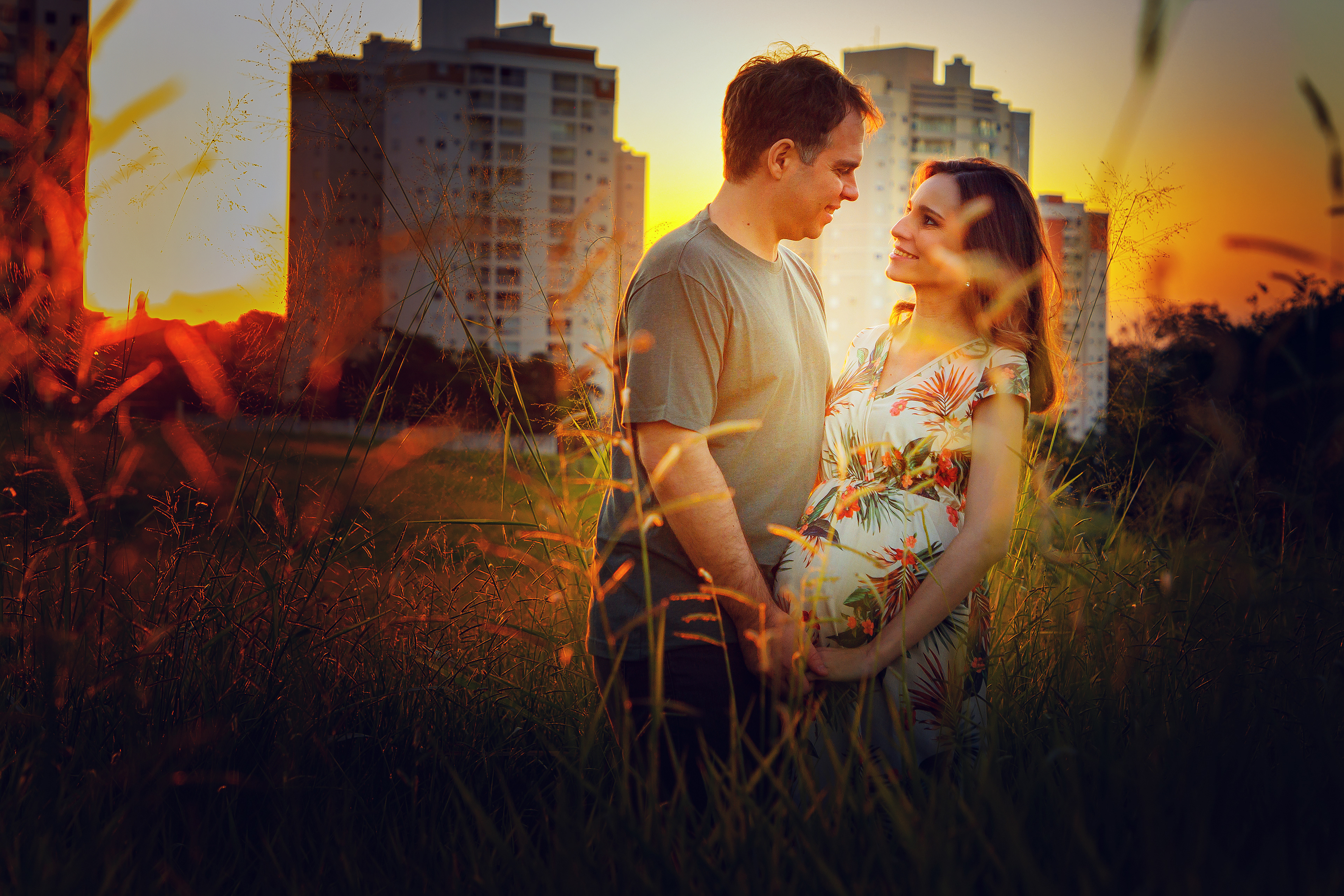 Foto dlm studio , ensaio externo de um casal em que a mulher esta gestante , uma troca de olhares e de maos dadas , estão entre a vegetação , predios ao fundo e uma linda luz dourada do por do sol