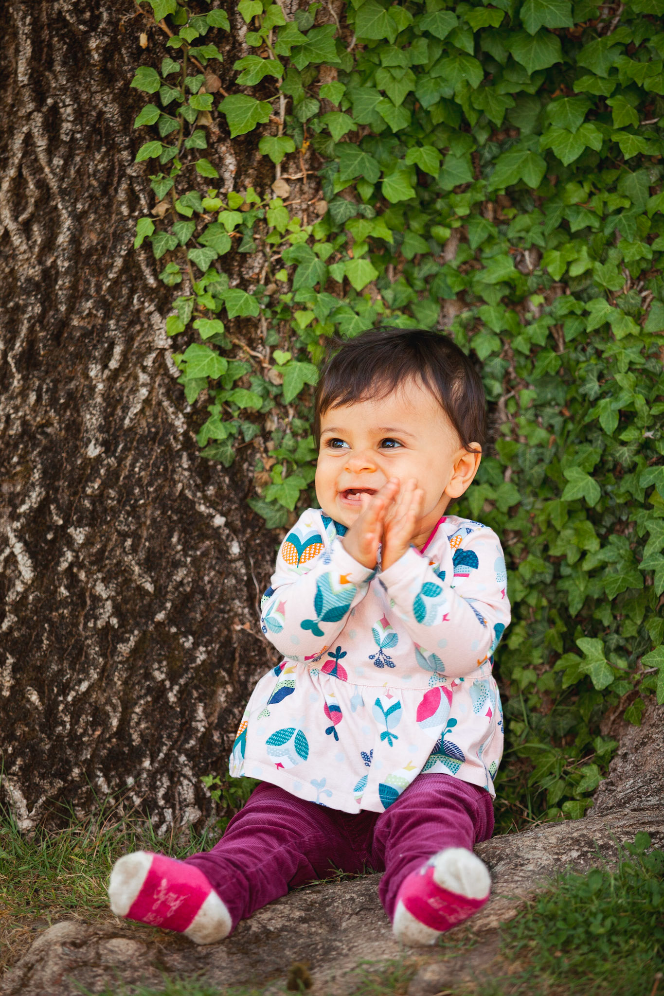 Petite fille assise contre un tronc d'arbre couvert de lierre, qui tape des mains. Photo couleur.