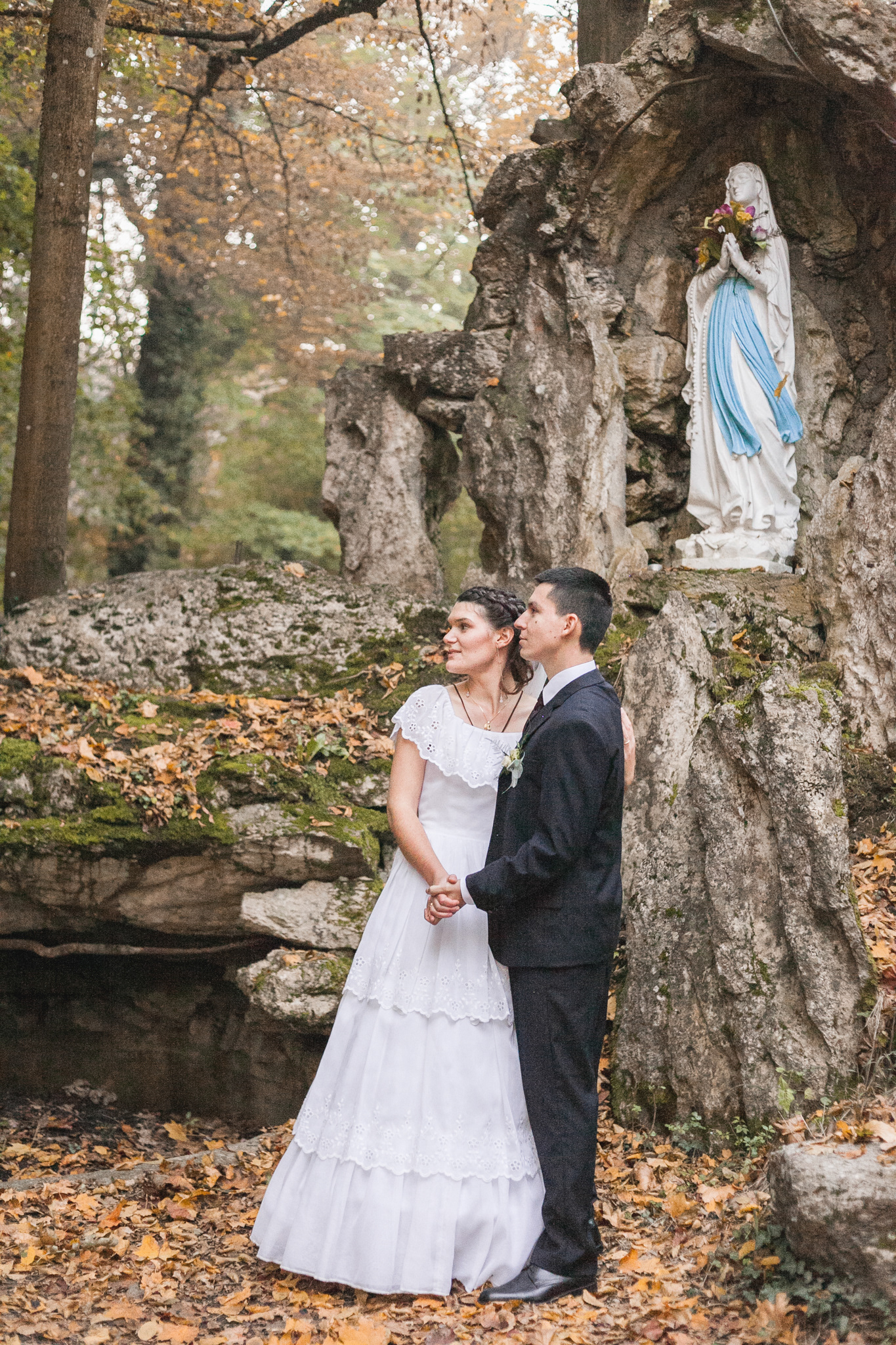 Couple de jeunes mariés enlacés sous une statue de la Vierge Marie. Photo couleur.