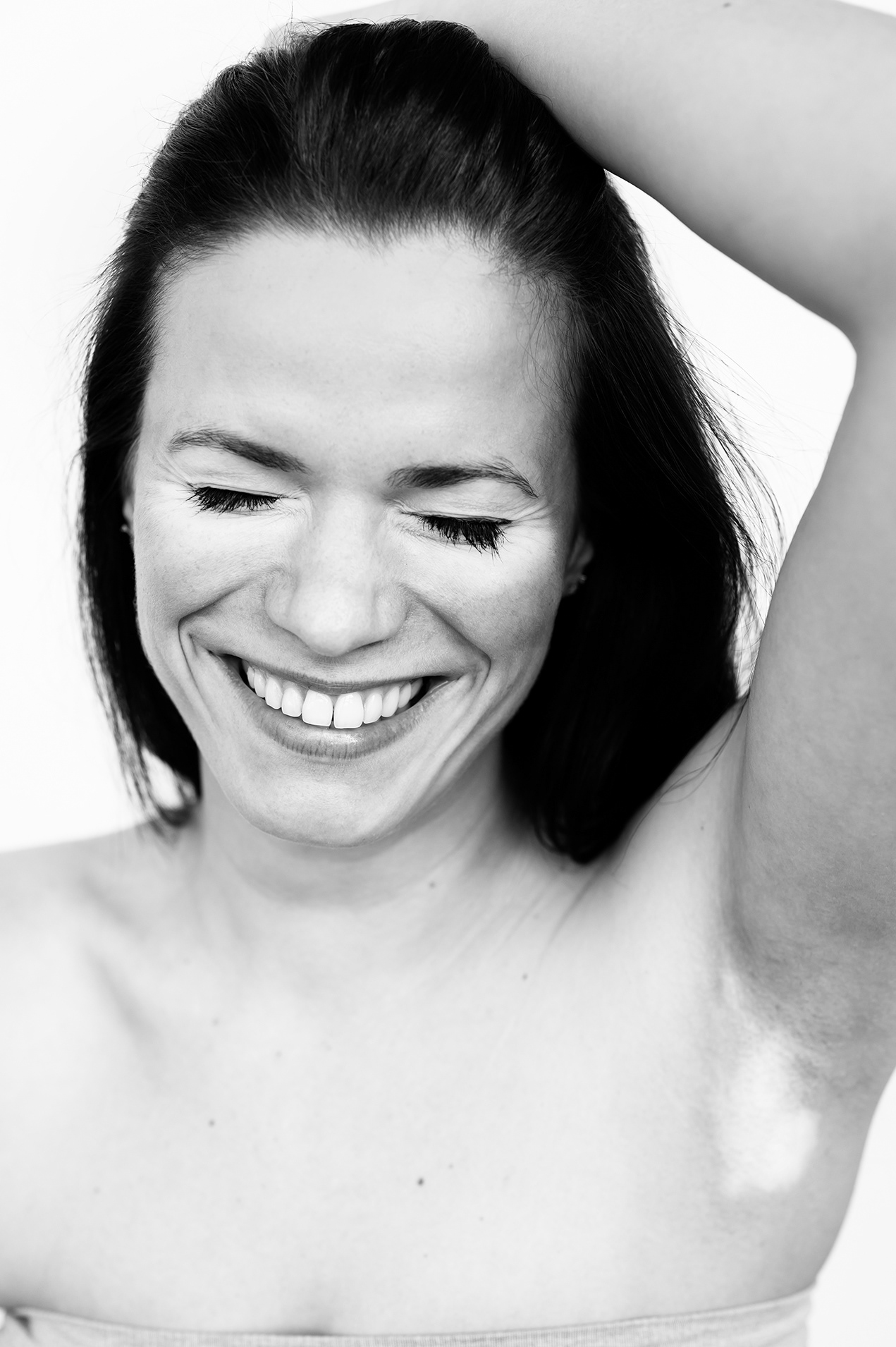 Black and white headshot. Mid-laugh, a model grins widely and shuts her eyes. She places 1 hand behind her head, revealing a single heart-shaped vitiligo patch on her armpit.