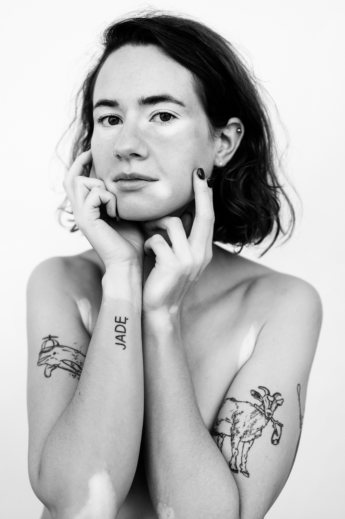 Black and white medium headshot. Contemplative and calm, the model lightly caresses down her jawline with both hands. Model has a light skin tone with several animal tattoos on her arms. Patches of vitiligo peek from her armpits, envelop her hands like gloves, and outline her eyes, mouth and elbows.